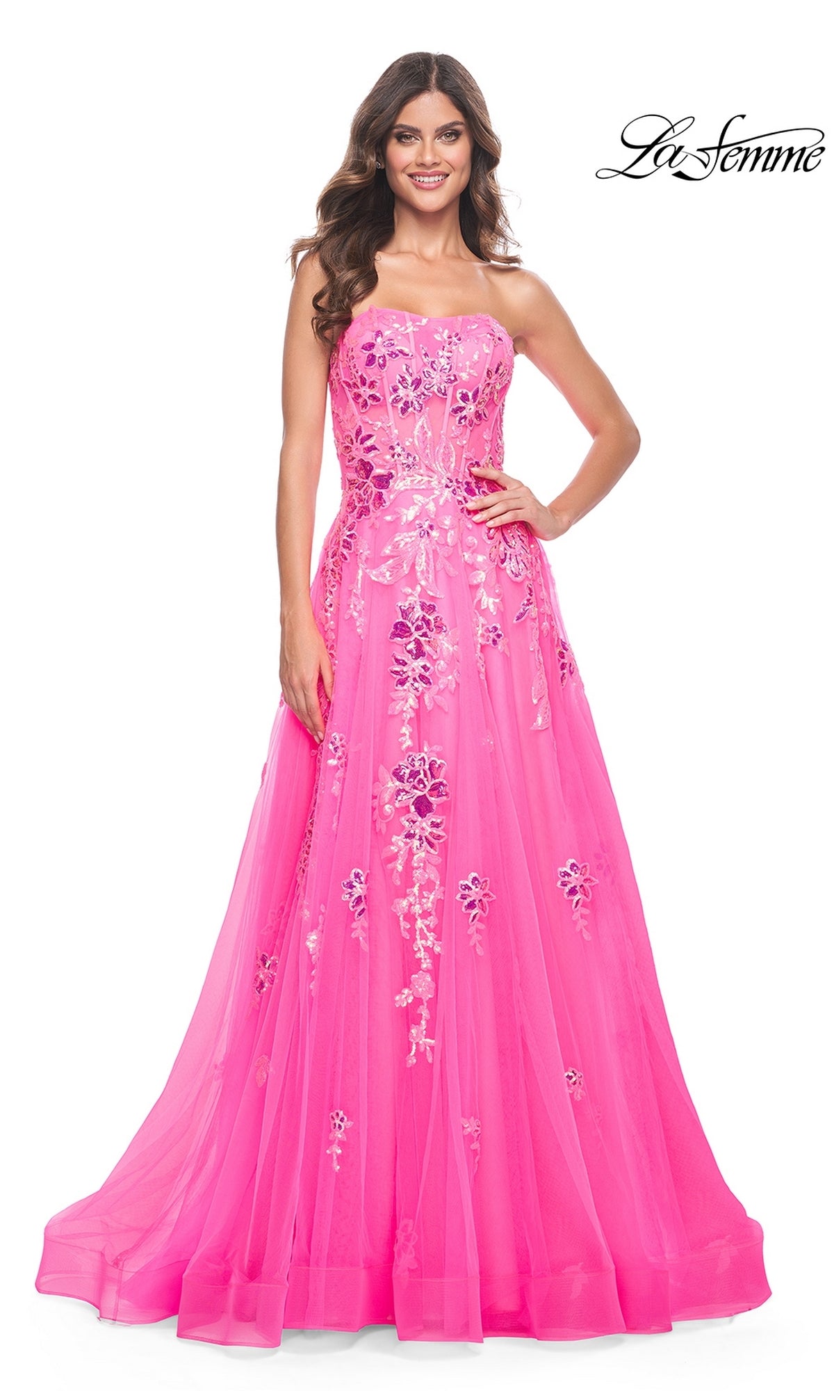 A woman wears the La Femme Neon Pink Long Prom Ball Gown 32137, featuring a strapless fitted bodice, sequin floral embellishments, and a full floor-length skirt. She stands against a white background, making a striking statement.