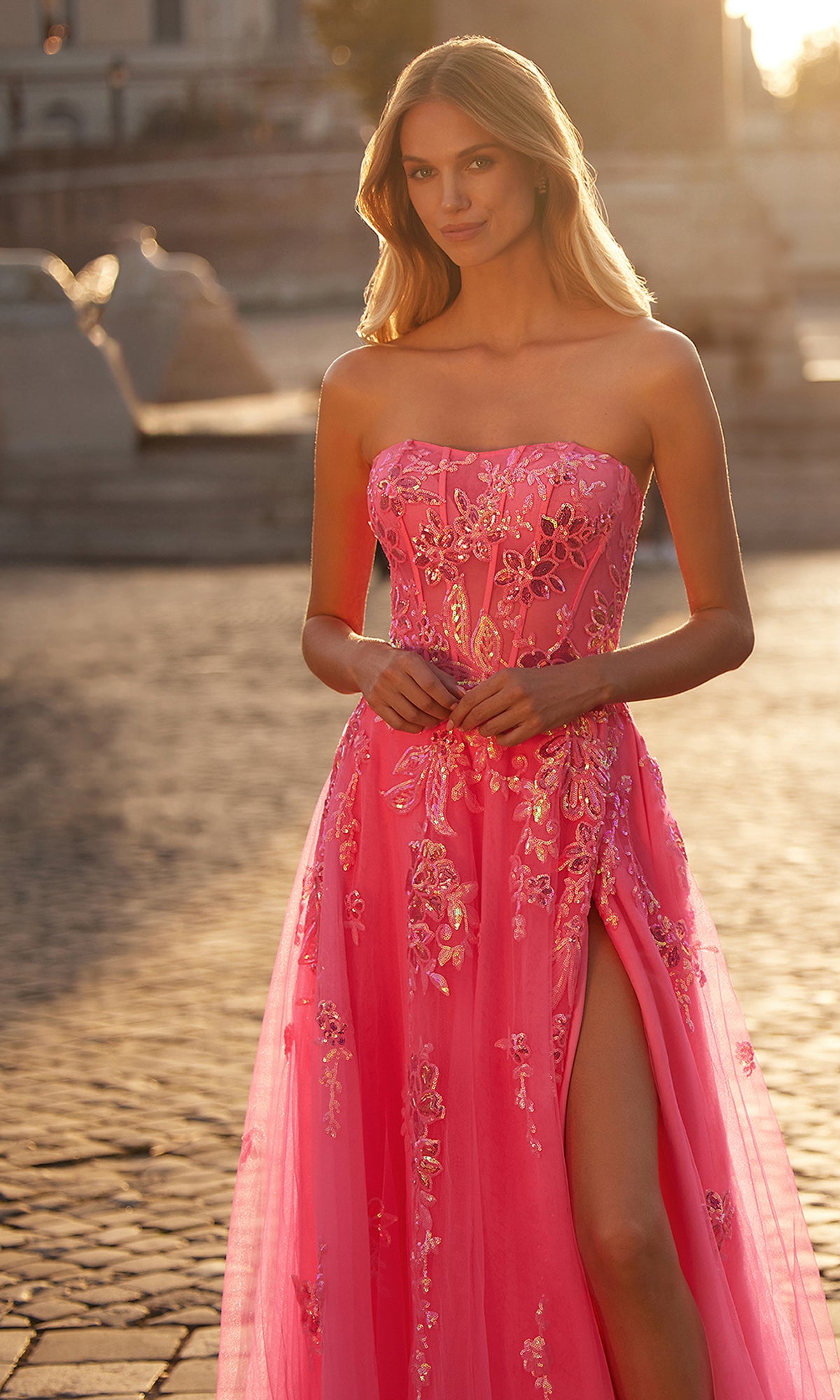 A woman stands outdoors on cobblestone pavement wearing the La Femme Neon Pink Long Prom Ball Gown 32137, featuring floral embroidery and a thigh-high slit.