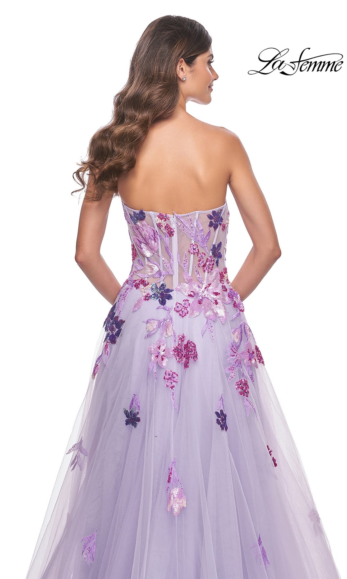 A woman with long, wavy hair models the La Femme Strapless Long Lavender Prom Dress 32156, featuring floral appliqués, a sheer bodice, and a flowing tulle skirt for an elegant, ethereal look—shown from the back.