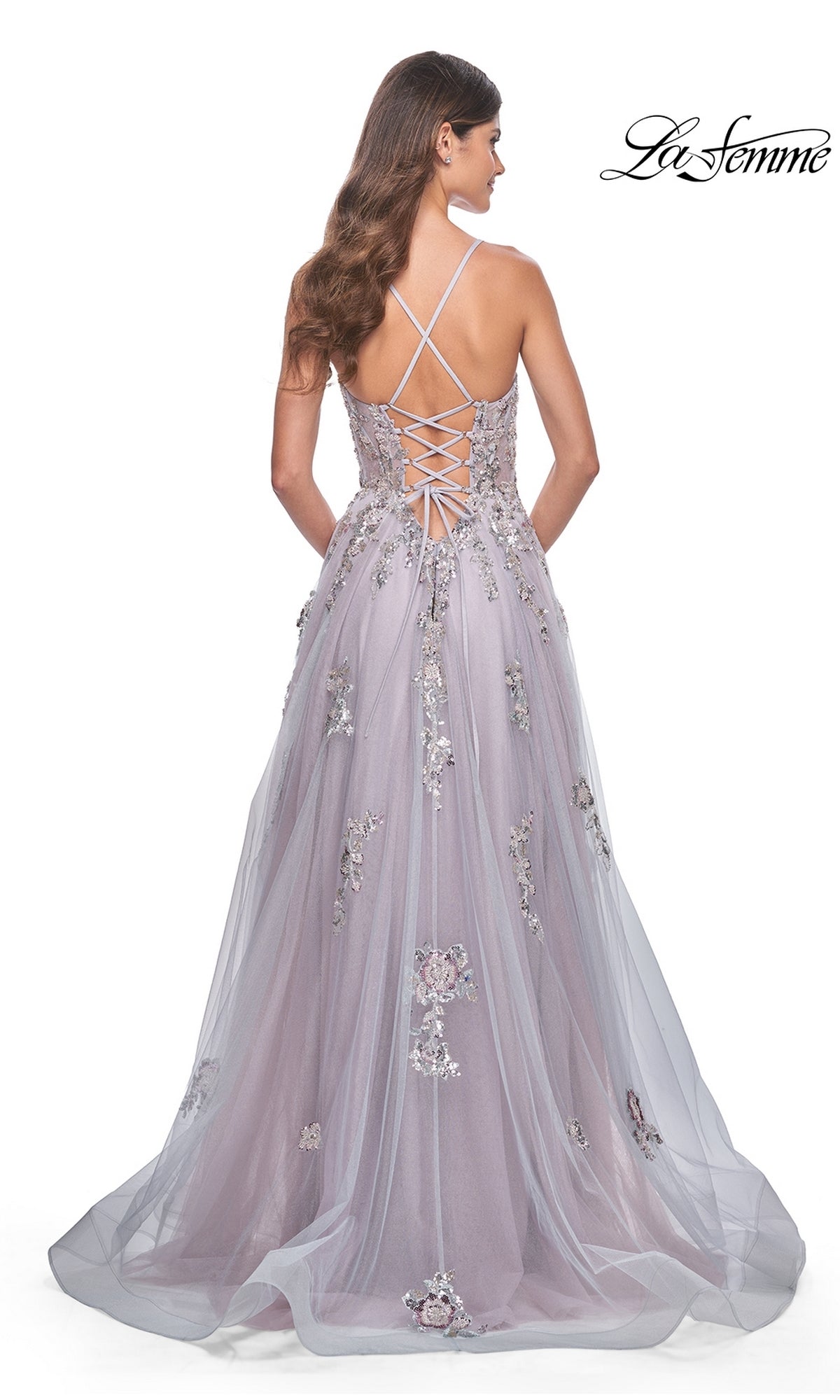A woman models the La Femme Strappy-Back Long A-Line Prom Dress 32200 in lavender, featuring floral embellishments, a lace-up back, and thin crisscross straps. La Femme logo appears in the top right corner.