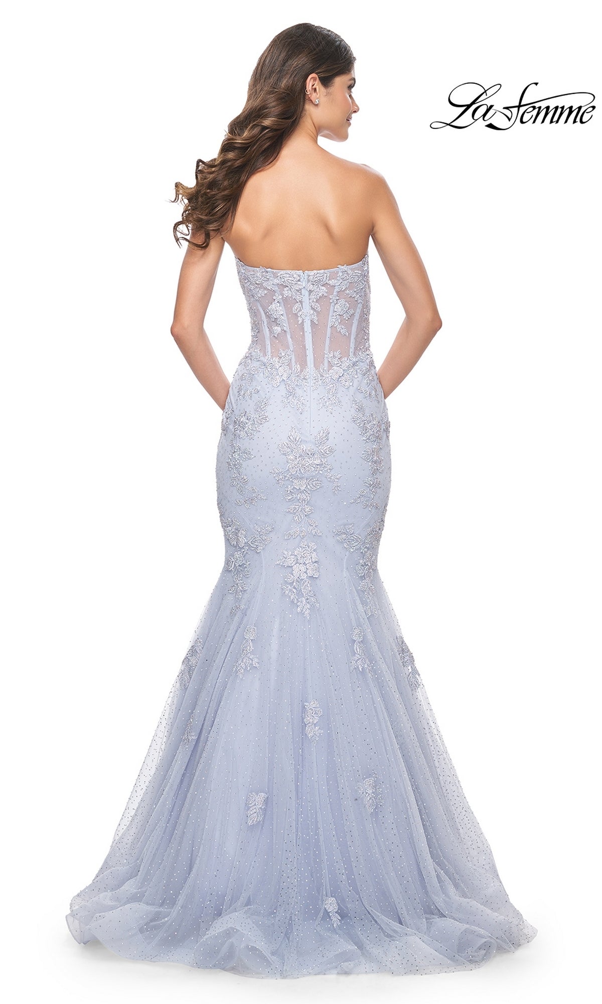 A woman wearing the La Femme Periwinkle Strapless Prom Dress 32214, featuring a fitted mermaid silhouette, sheer embroidered details, and a tulle train, viewed from the back.