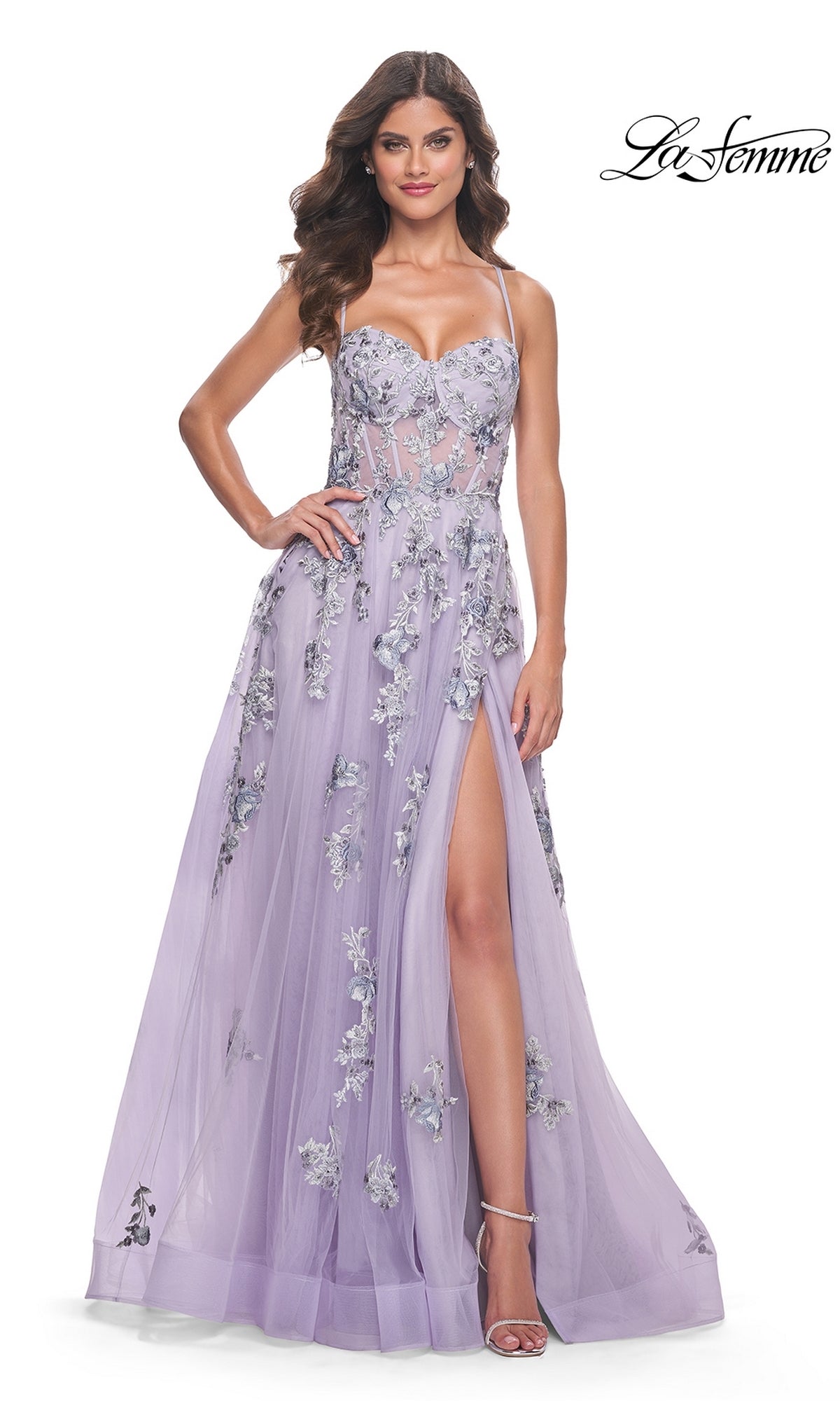 A woman models the La Femme Long Lavender A-Line Prom Dress 32221, featuring floral appliqué, a sheer bodice, thin straps, and a high side slit. She poses elegantly with one hand on her hip. The