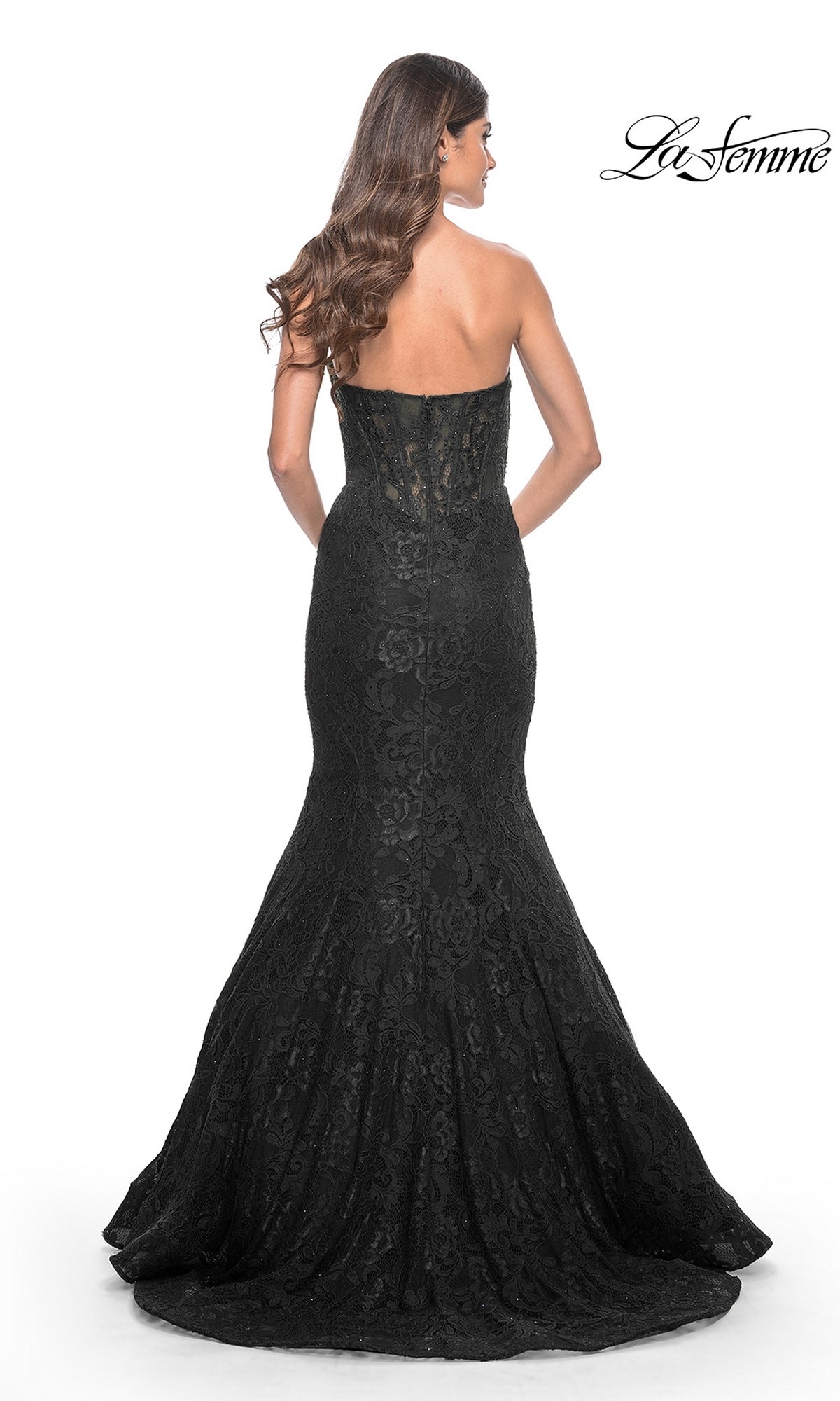 A woman wearing the La Femme Strapless Long Mermaid Prom Dress 32249 in black stands facing away, her long wavy hair flowing down her back. The fitted bodice and flared skirt emphasize the elegant lace details of this floor-length gown.
