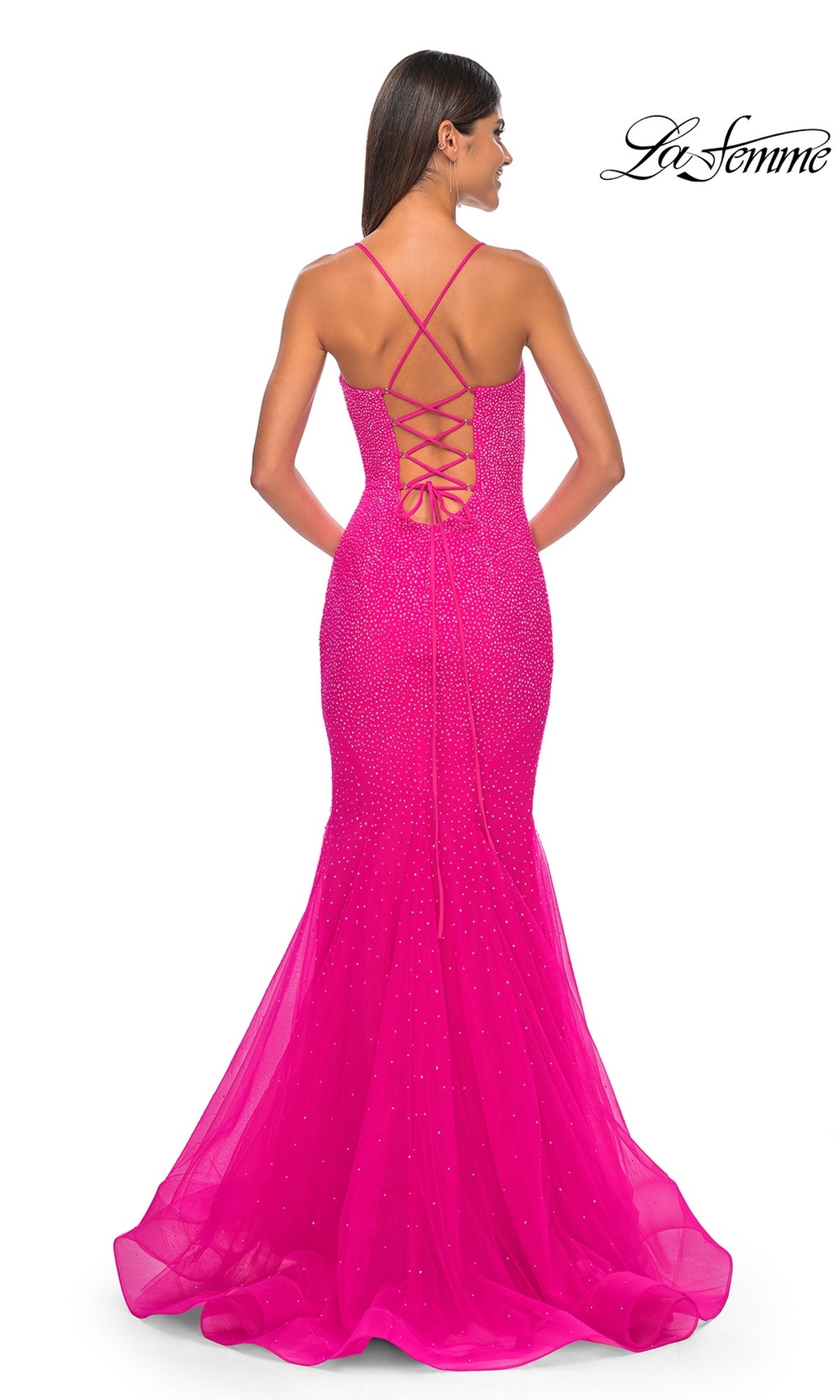 A woman faces away from the camera in the La Femme Embellished Long Mermaid Prom Dress 32273, a bright pink floor-length gown with a lace-up open back and thin crisscross straps.