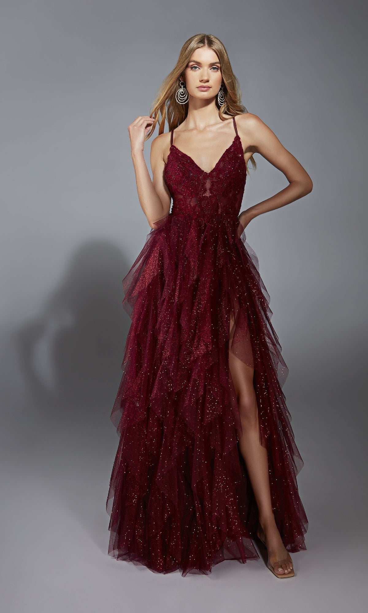 A woman poses against a gray background wearing the Aerie Couture 322 Glitter Ruffle Prom Ball Gown—a sleeveless, floor-length dress with a v-neck lace bodice, sparkling details, and a thigh-high leg slit.