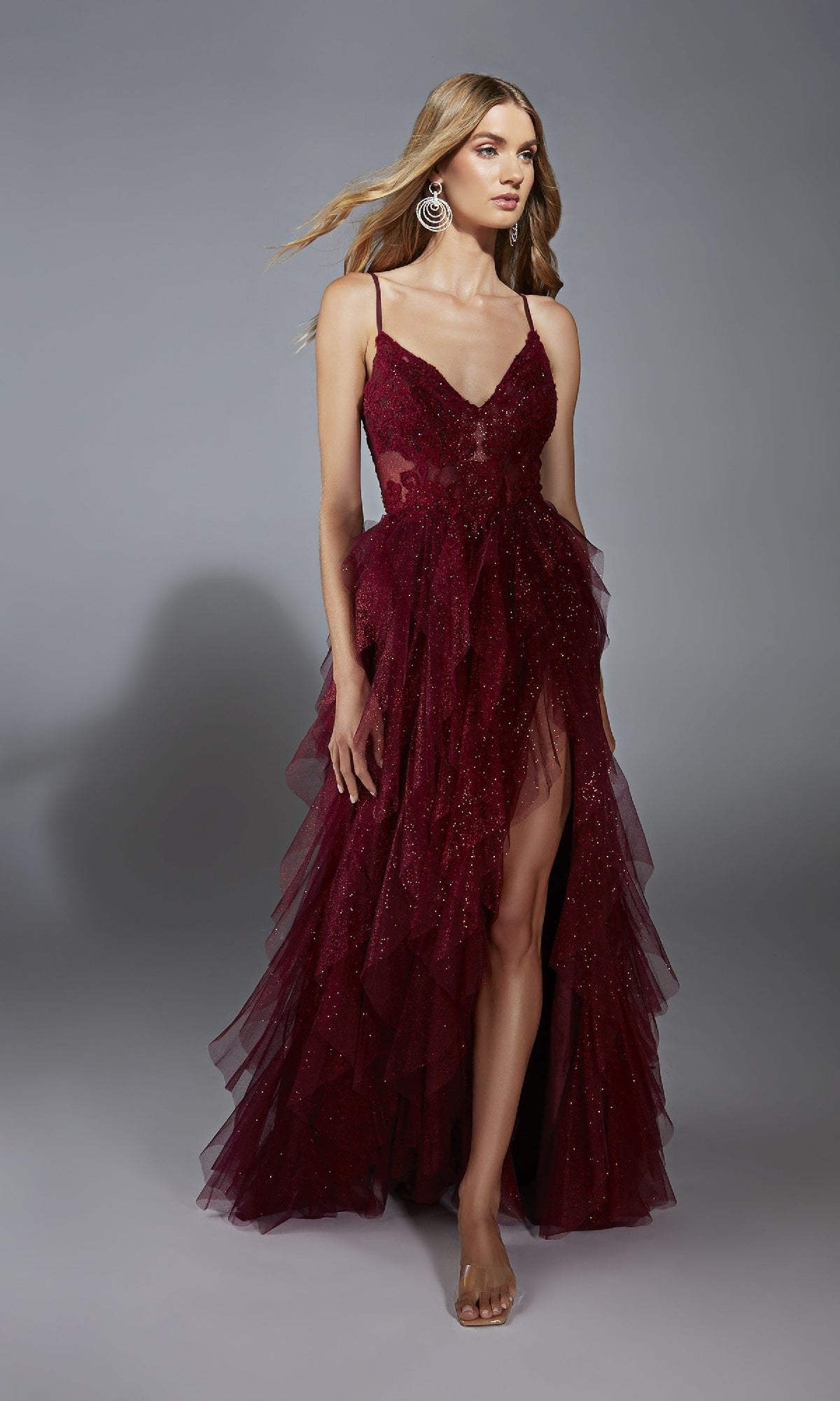 A woman wears the Aerie Couture 322 Glitter Ruffle Prom Ball Gown, a sleeveless deep burgundy dress with sparkly embellishments, high leg slit, thin straps, and layered tulle skirt, posing against a plain gray background.