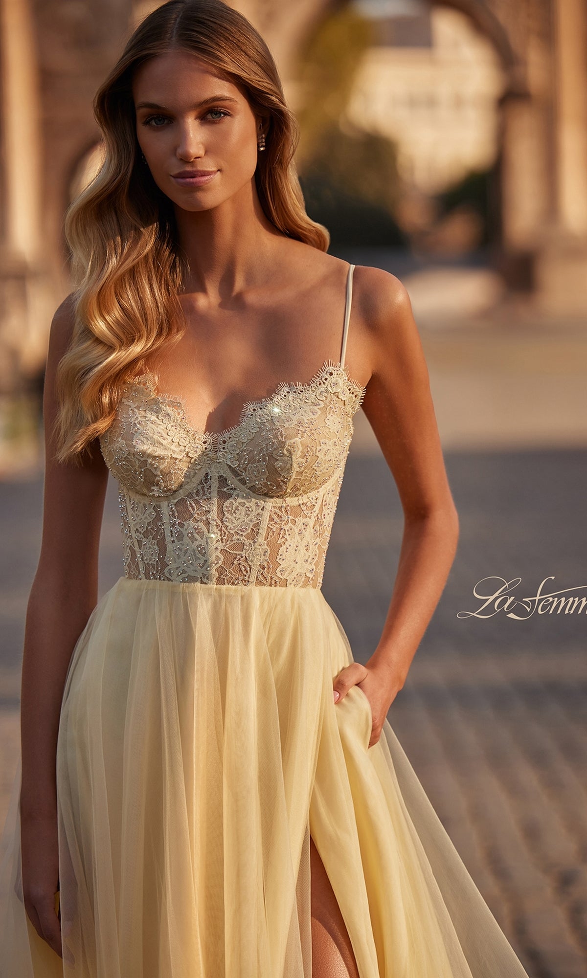 A woman in a sheer yellow La Femme Lace-Bodice Long A-Line Prom Dress 32306 stands outdoors on a cobblestone path, with an arched stone structure in the background.