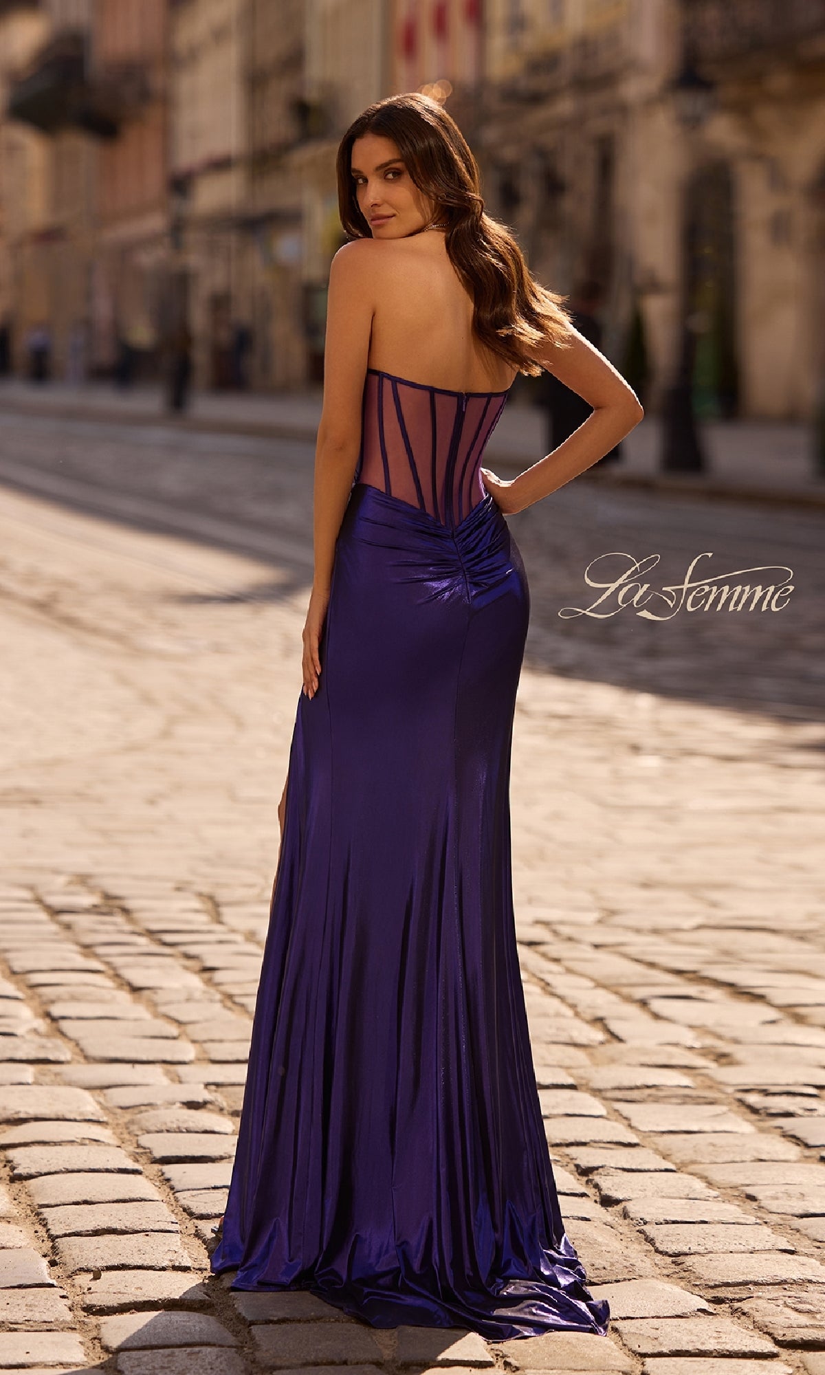 A woman stands on a cobblestone street, facing away, wearing the Strapless Long Metallic Prom Dress La Femme 32375 in deep blue satin with a sheer corset back. The