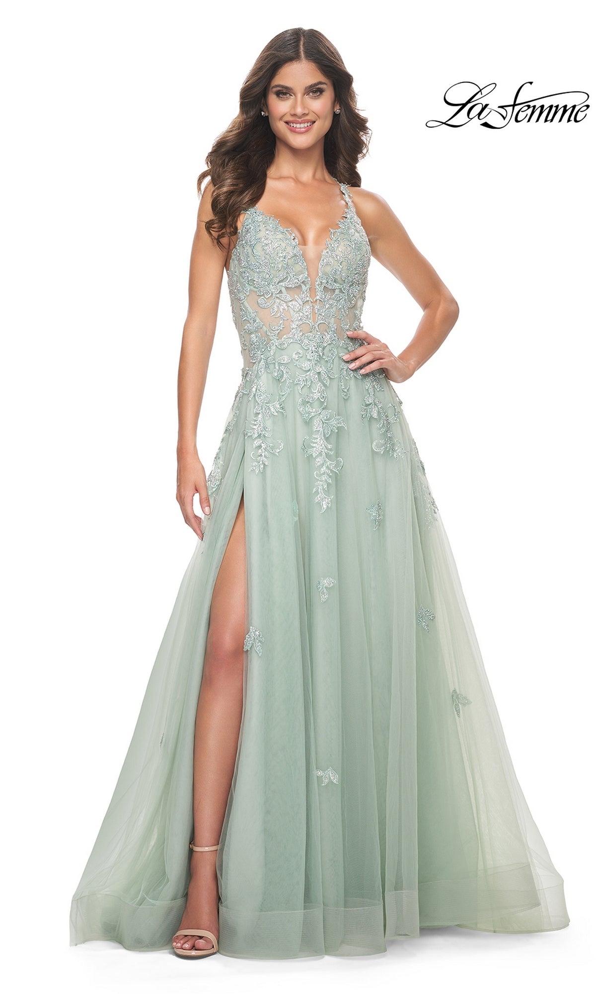 A woman stands smiling at the camera wearing the Sheer-Waist Long A-Line Prom Dress: La Femme 32438, a light green formal gown featuring floral embroidered lace, sleeveless design, deep V neckline, sheer bodice, and a high slit.