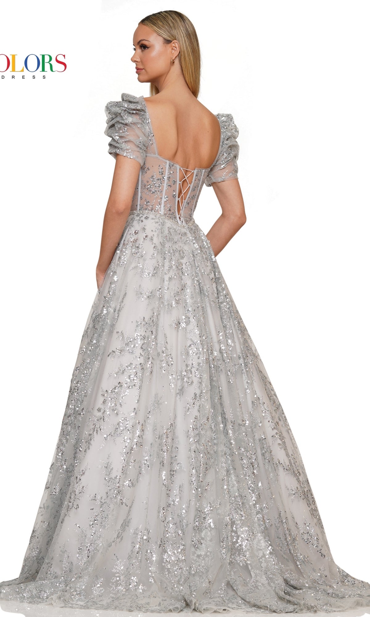 A woman stands facing away, wearing the Short-Sleeve Long Glitter A-Line Prom Dress 3243, featuring a silver floor-length design with puffed sleeves, sequin accents, and a lace-up back.