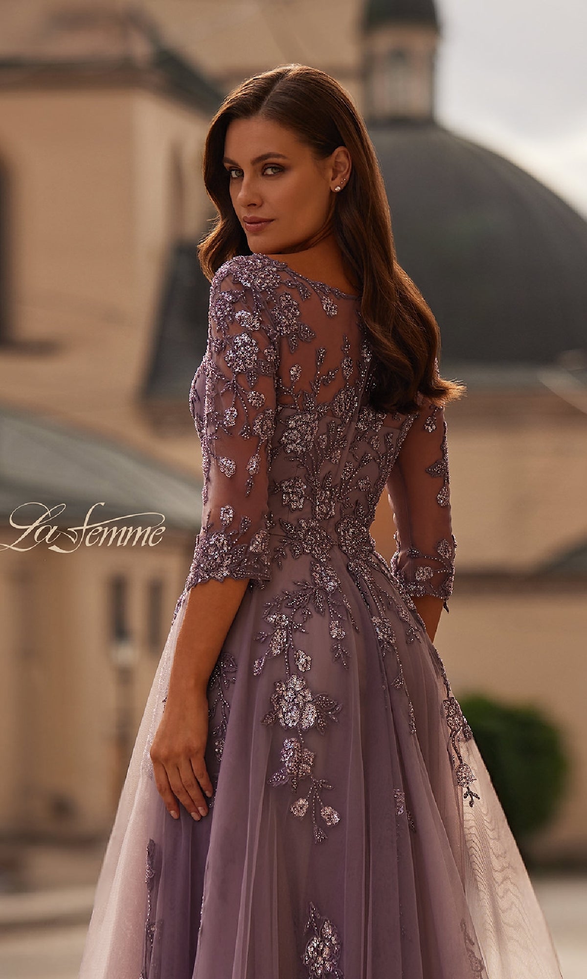 A woman wearing the La Femme 32538 A-Line Tulle Sleeved Formal Dress, featuring lavender floral embroidery and long sleeves, stands outdoors looking over her shoulder.