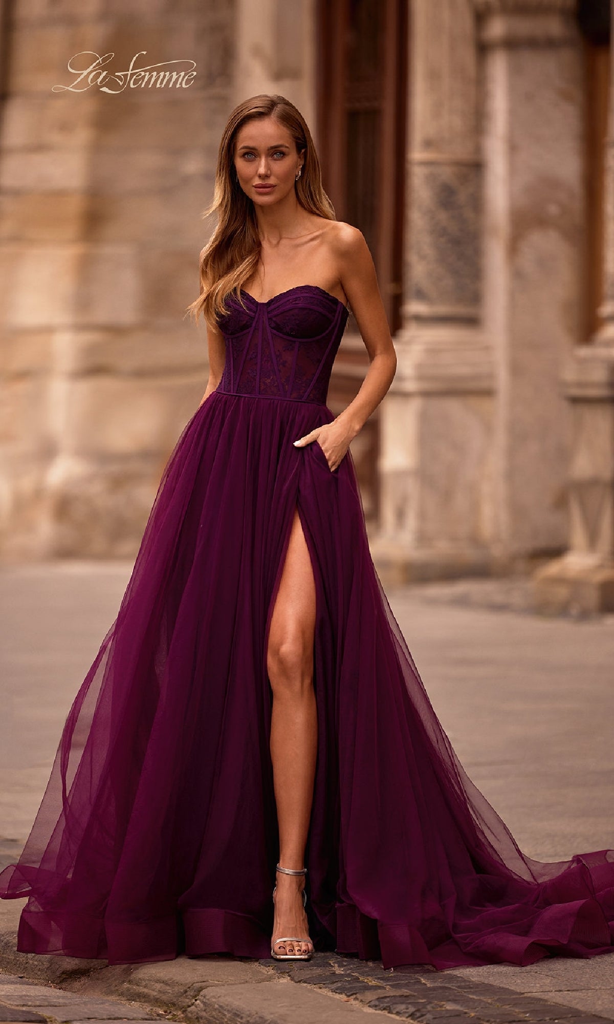 A woman wearing the La Femme 32579 Strapless Long Prom Ball Gown, featuring a floral corset-style bodice and thigh-high slit, stands outside on stone pavement in front of a historic building.