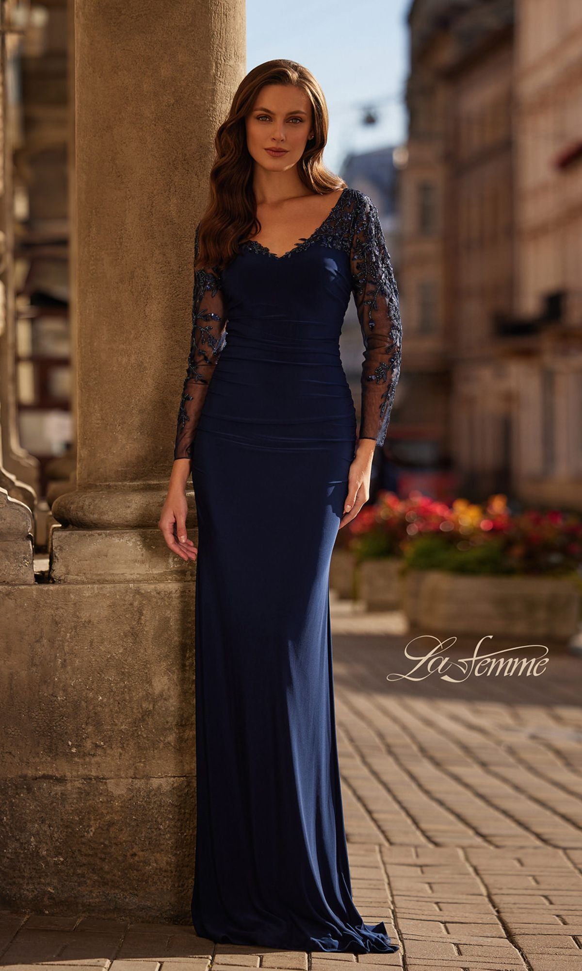 A woman stands outdoors in the La Femme 32609—a long navy formal dress with sheer, beaded long sleeves—set against elegant stone columns and buildings.