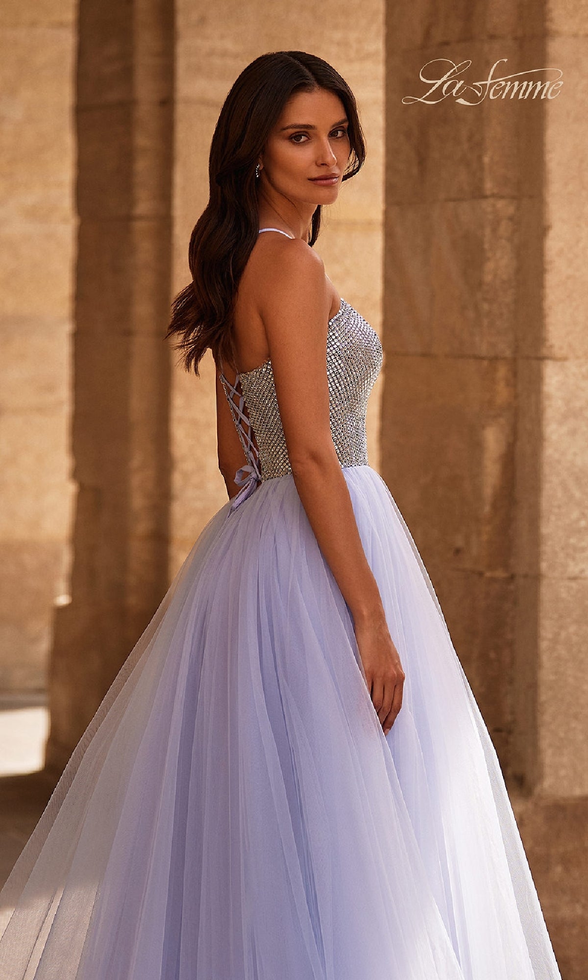 A woman models the Beaded-Corset Periwinkle Prom Gown: La Femme 32633, standing sideways before stone columns in a dress featuring a rhinestone-beaded corset back.