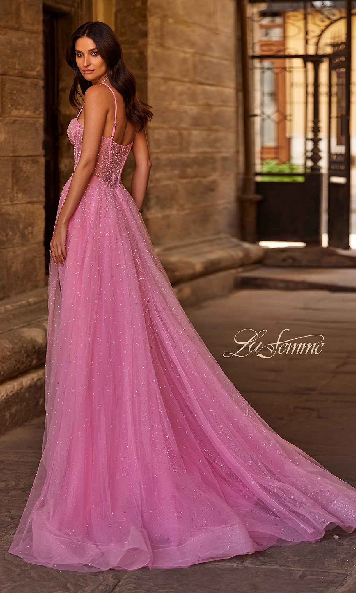 A woman wearing the Pearl Classic Long Prom Dress: La Femme 32644 stands outdoors in front of a stone building with wrought iron gates, showcasing the gown's fitted bodice and flowing train.