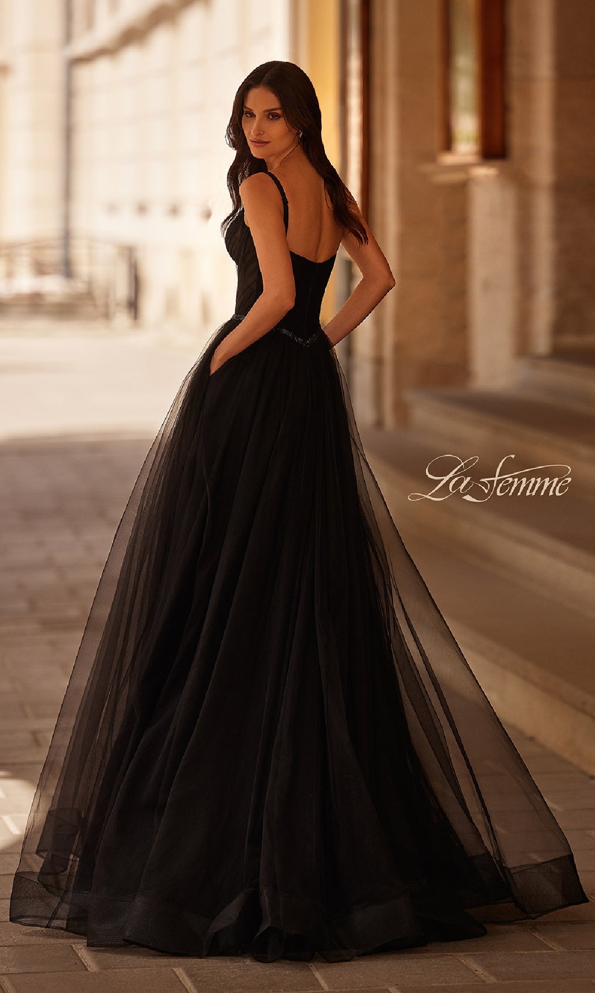 A woman stands outdoors, looking over her shoulder in the La Femme 32956 Beaded-Waist Long Prom Ball Gown, featuring a black full tulle skirt and elegant corset bodice.