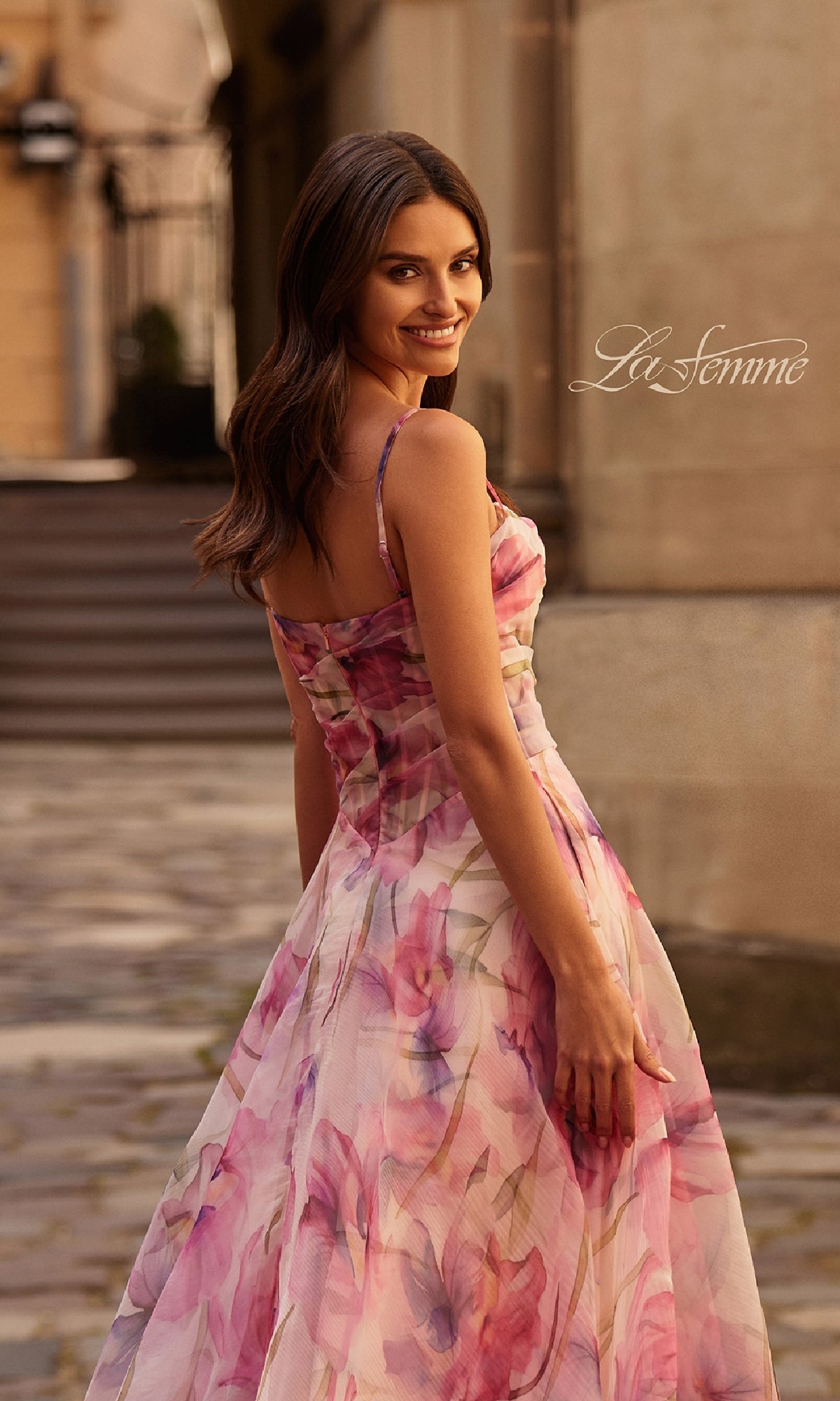 A woman wearing the Pink Floral-Print Corset Prom Dress: La Femme 32686 stands outdoors on a stone path, looking back over her shoulder and smiling. The