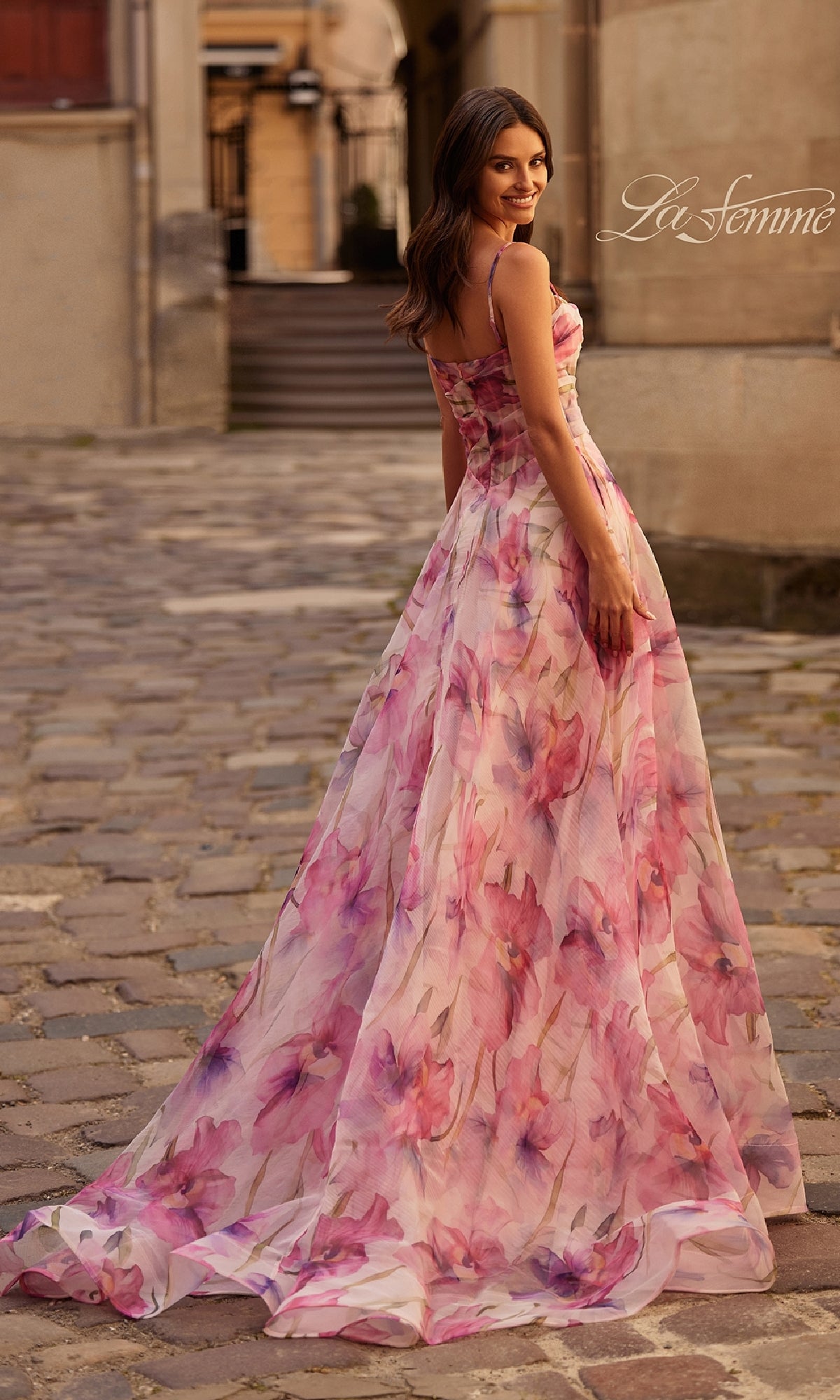 A woman wearing the La Femme 32686 Pink Floral-Print Corset Prom Dress with thin straps and a subtle train stands on a cobblestone street, glancing back over her shoulder.