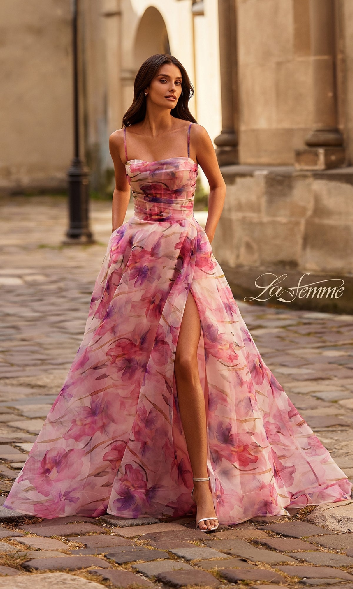 Woman wearing the La Femme 32686 Pink Floral-Print Corset Prom Dress with a high slit, standing on a cobblestone street with stone buildings in the background.