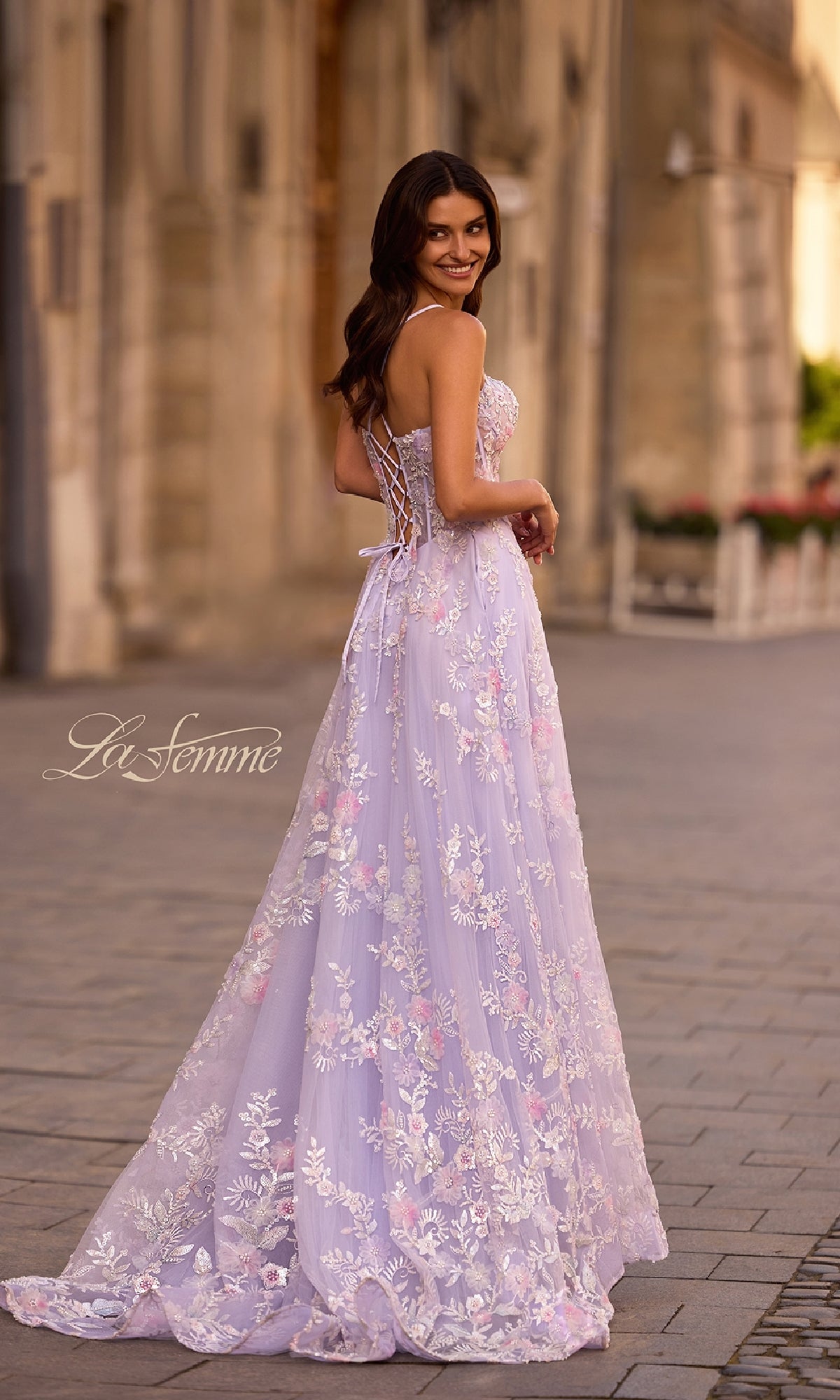 Wearing the La Femme 32695 long prom dress in light purple with floral lace embroidery, a woman stands on a cobblestone street, looking over her shoulder and smiling.