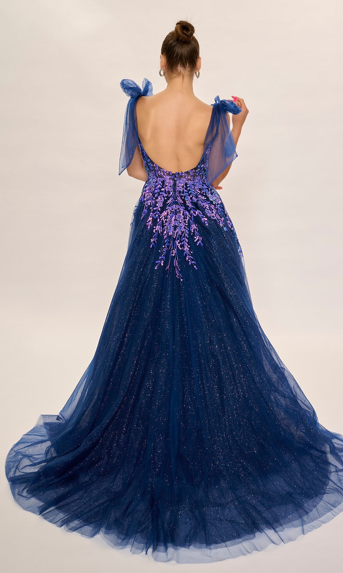 A woman in a dark blue Nina Canacci 3269 long formal dress featuring a low back, purple floral details, tulle overlay, embellished fitted bodice, and bow straps stands against a plain background.