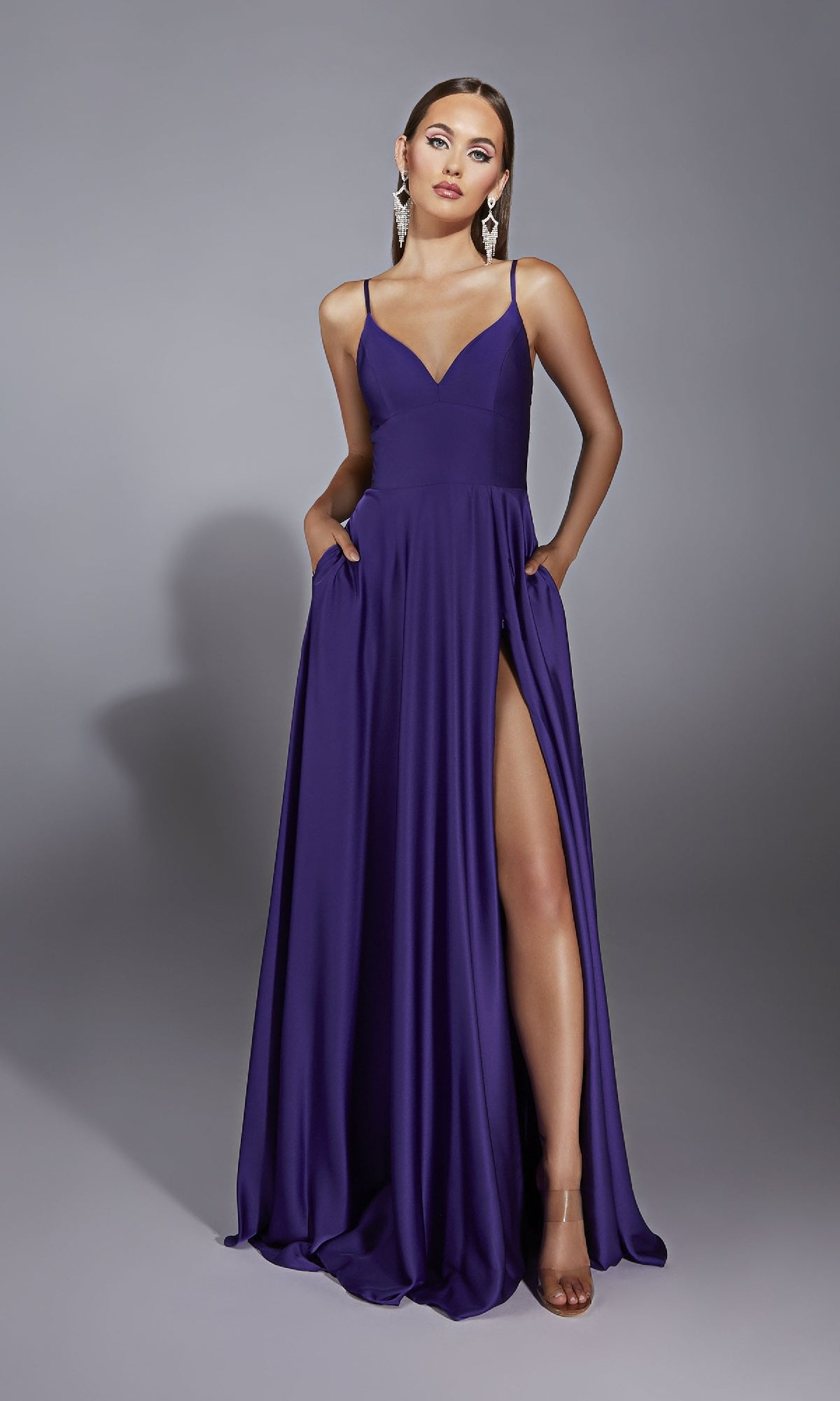 A woman wears the Long Formal Dress: Aerie Couture 326—a sleeveless, floor-length purple a-line prom dress with a high slit and pockets—paired with dangling earrings and open-toe heels, against a plain gray background.