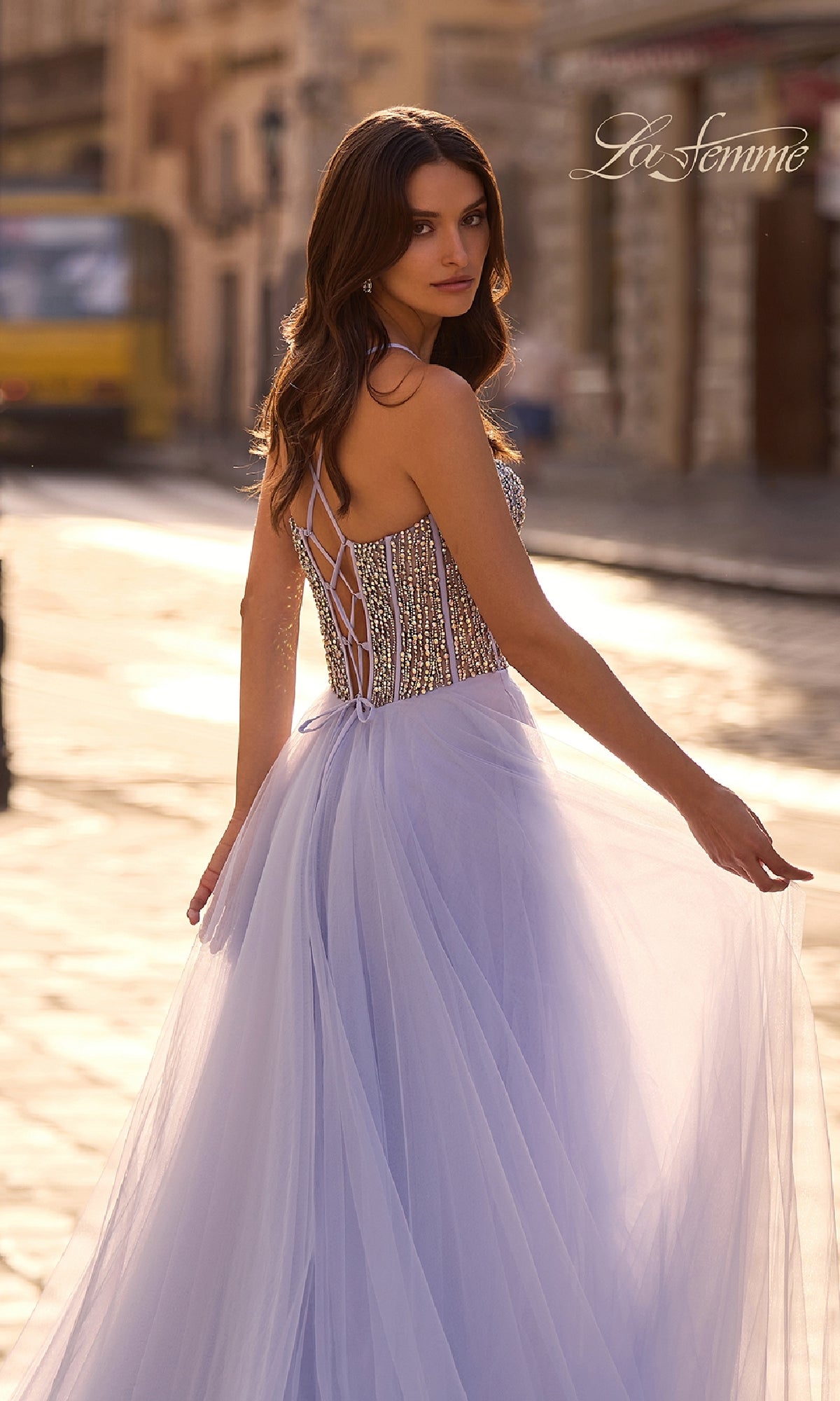 A woman wearing the La Femme 32703 Beaded-Corset Long Prom Dress, a sleeveless lavender A-line gown with a lace-up back and rhinestone corset, stands on a sunlit city street looking over her shoulder.