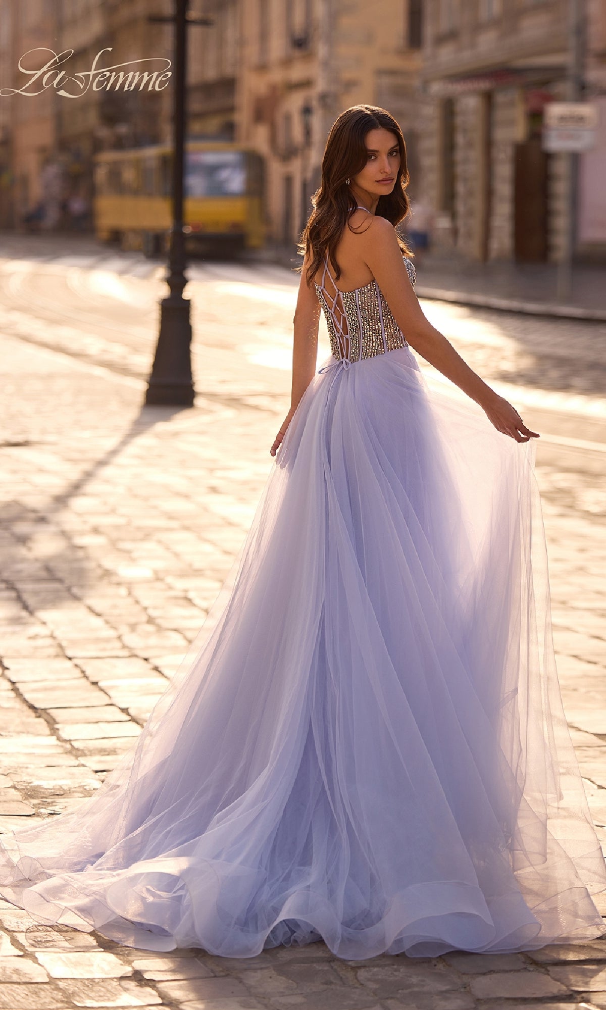 Wearing the La Femme 32703 Beaded-Corset Long Prom Dress in light blue, a woman stands on a cobblestone street and glances back over her shoulder as a yellow tram passes by.