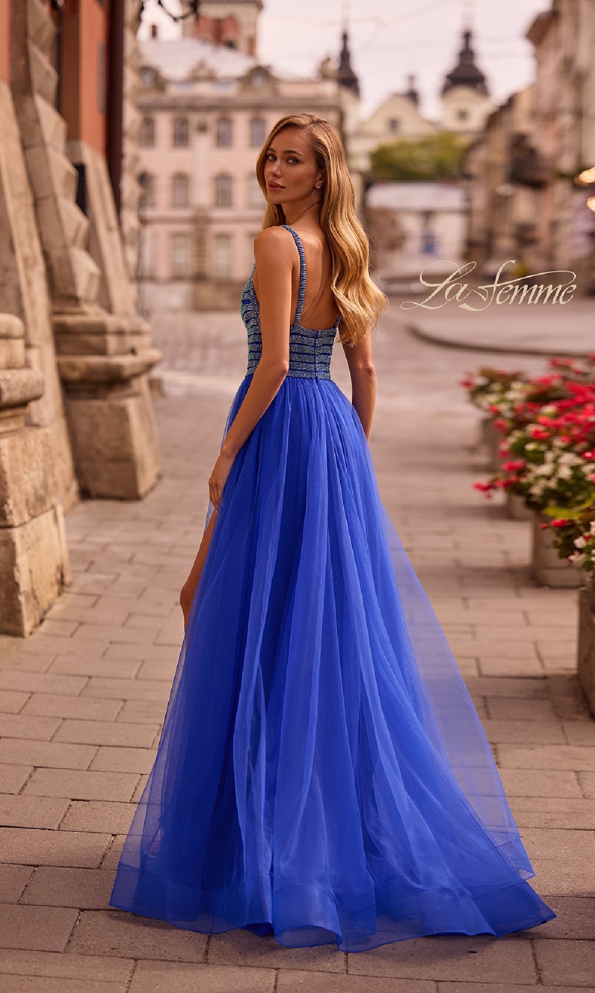A woman in the Rhinestone-Bodice Prom Ball Gown: La Femme 32714 stands on a stone street, looking over her shoulder, with flowers and historic buildings in the background.