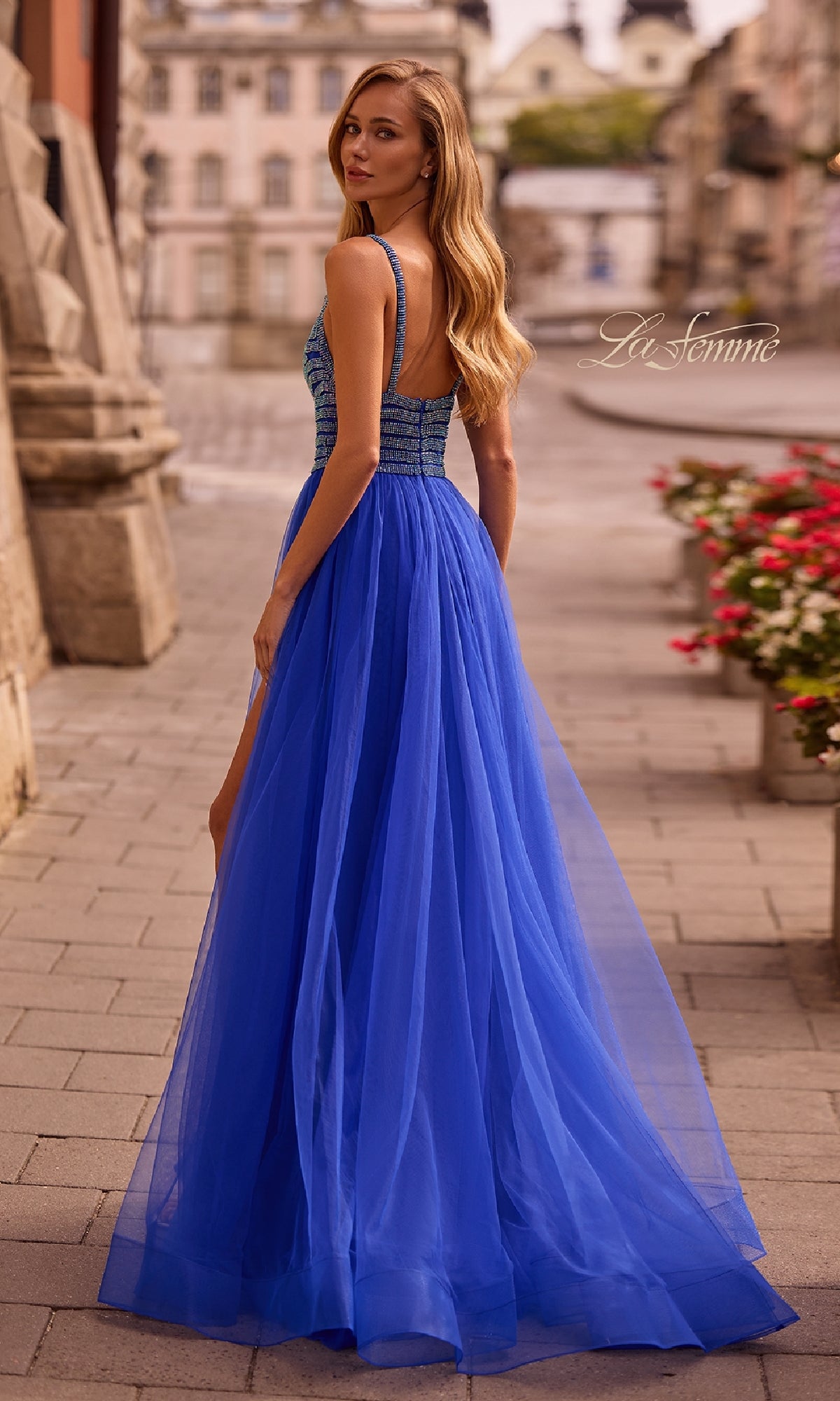 A woman in the La Femme 32714 Rhinestone-Bodice Prom Ball Gown poses on a cobblestone street with stone buildings and potted flowers, glancing over her shoulder.