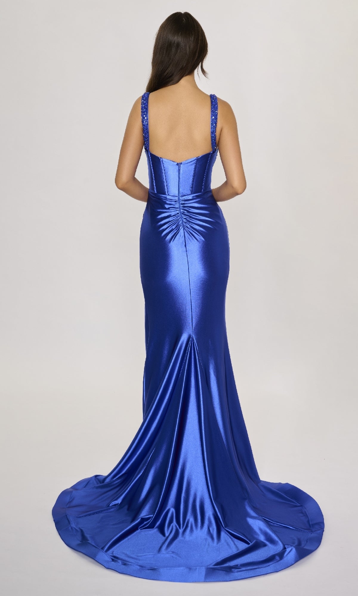A woman with long dark hair is seen from the back wearing the Nina Canacci 3271 long formal royal blue satin dress, featuring a fitted corset bodice and floor-length train, set against a plain light background.