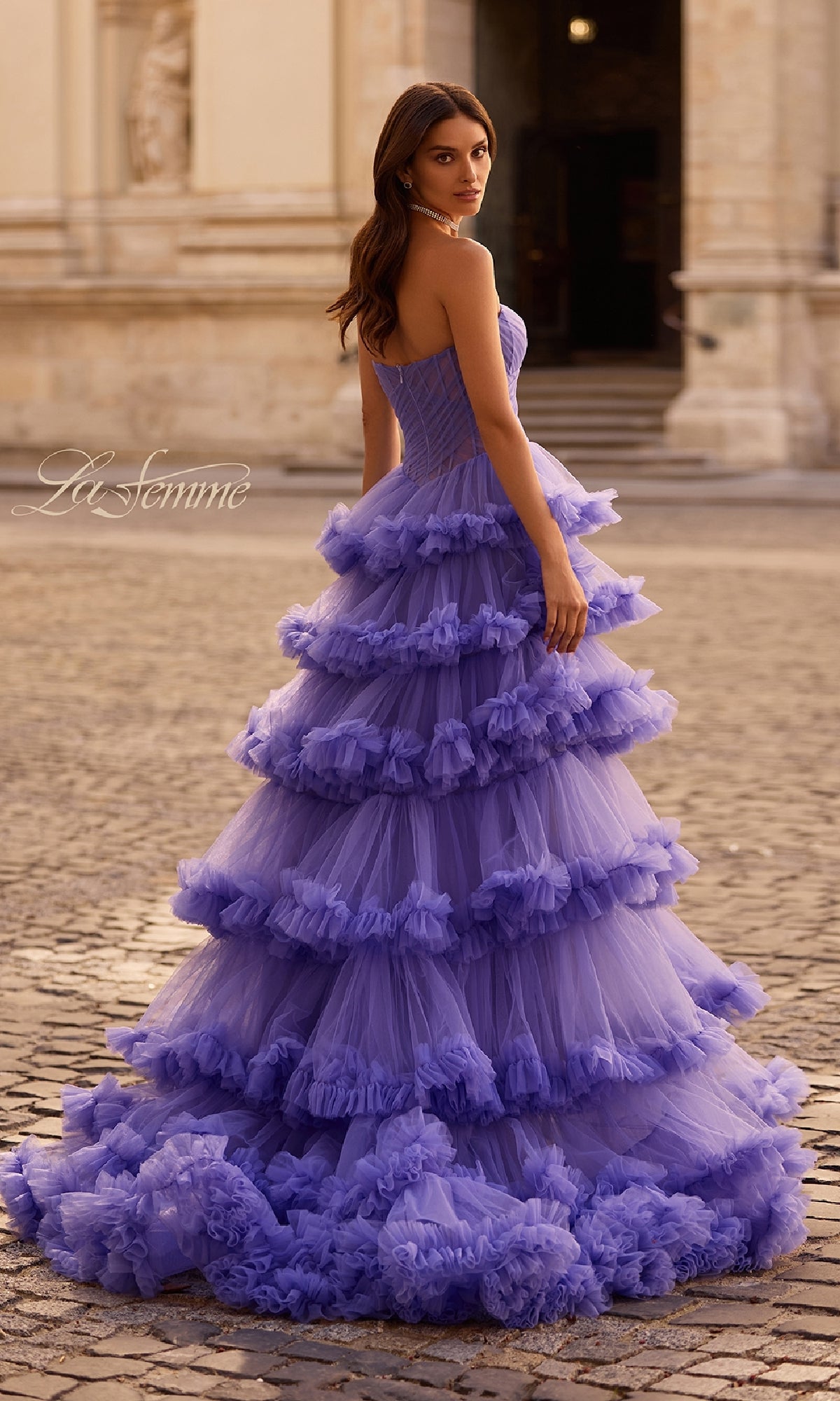 A woman outdoors wears the Strapless Ruffled Prom Ball Gown: La Femme 32721—a long, tiered lavender dress with ruffles—glancing back over her shoulder.