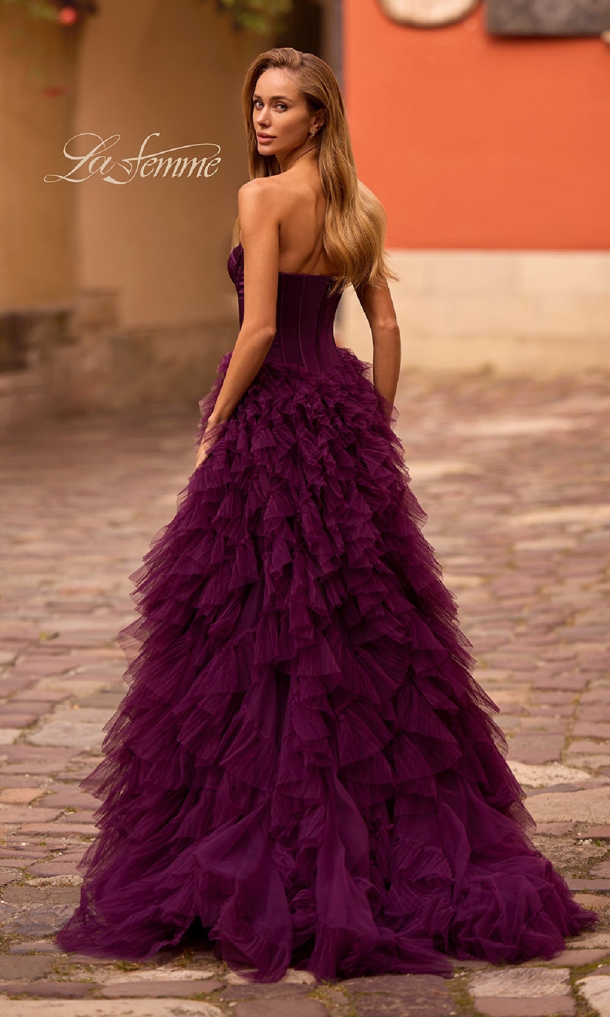 A woman in the La Femme 32760 Strapless Long Ruffled Prom Dress with a corset-style bodice stands on a cobblestone street, glancing back over her shoulder.