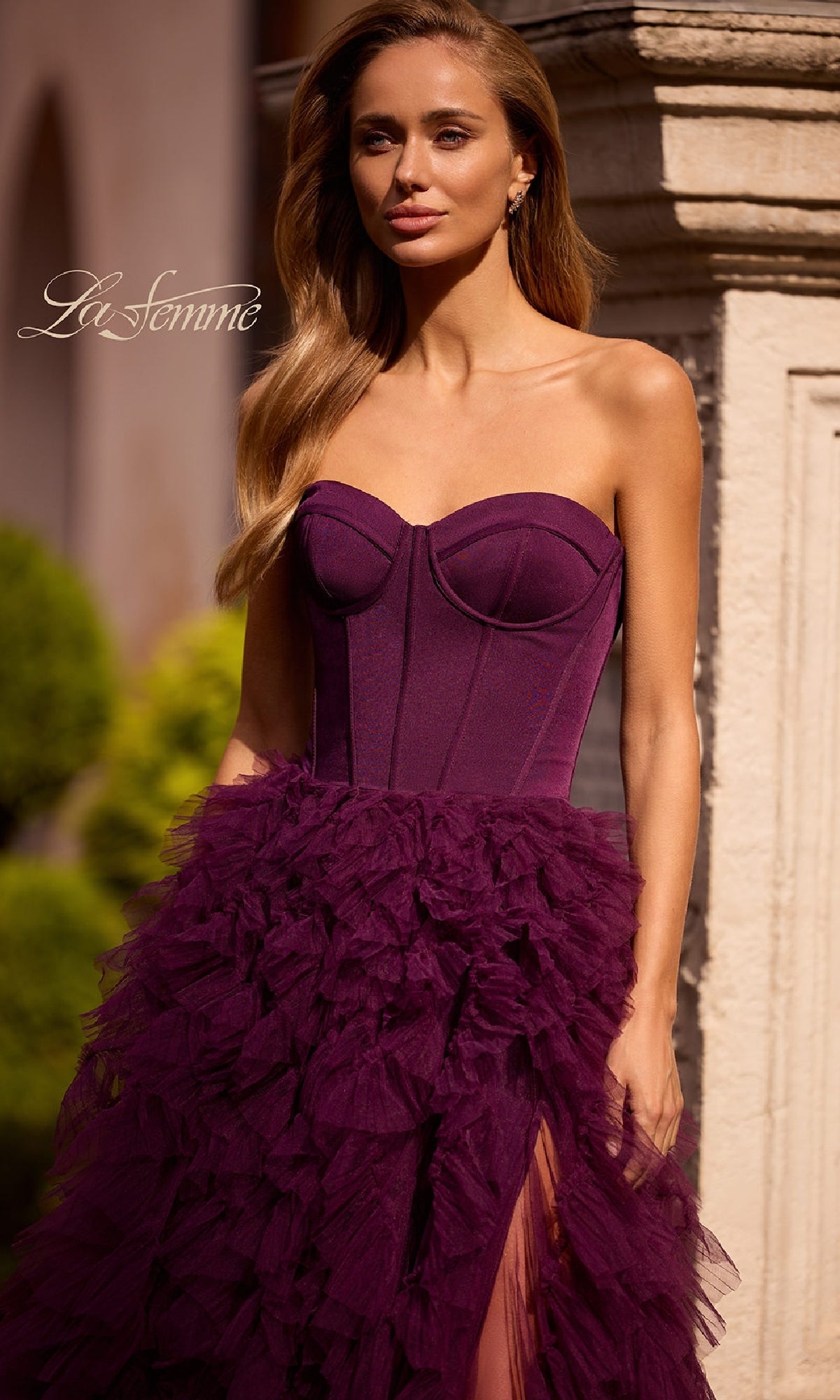 A woman stands outdoors near a stone column wearing the La Femme 32760, a strapless long ruffled prom dress in deep purple featuring a corset-style bodice and layered tulle A-line skirt.