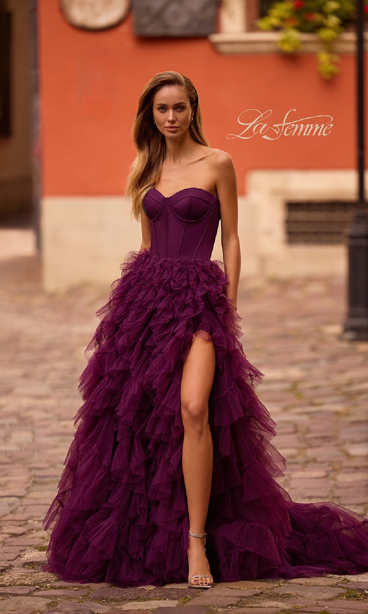 A woman stands outdoors on a cobblestone street, wearing the La Femme 32760 Strapless Long Ruffled Prom Dress with a thigh-high slit and corset-style bodice.