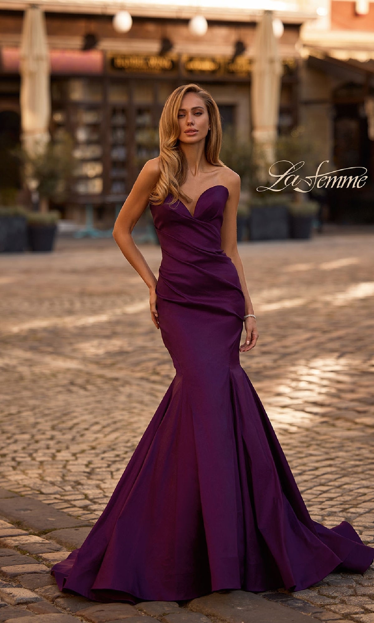 A woman stands on a cobblestone street in the Strapless Long Mermaid Prom Dress: La Femme 32791, a dark purple gown, with storefronts and outdoor seating visible in the background.