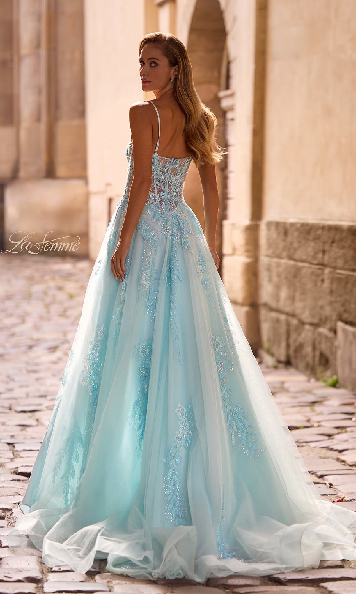 A woman in the La Femme 32810 long prom dress, featuring light blue tulle, beaded lace appliqué, and thin straps, stands on a cobblestone path looking over her shoulder.