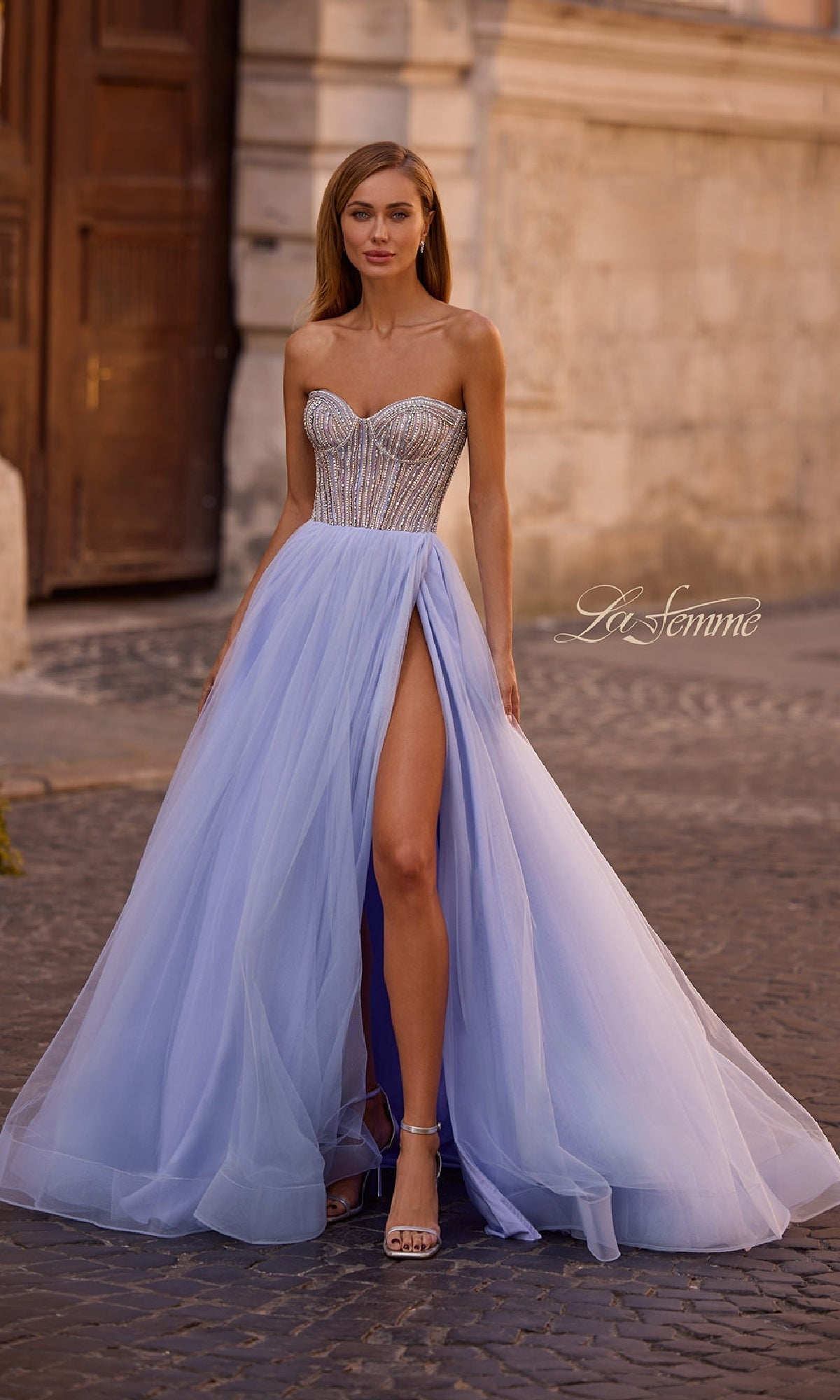 A woman poses outdoors in front of a stone building, wearing the La Femme 32811 Strapless Long Prom Ball Gown featuring a lavender hue, beaded sweetheart corset, and high slit.