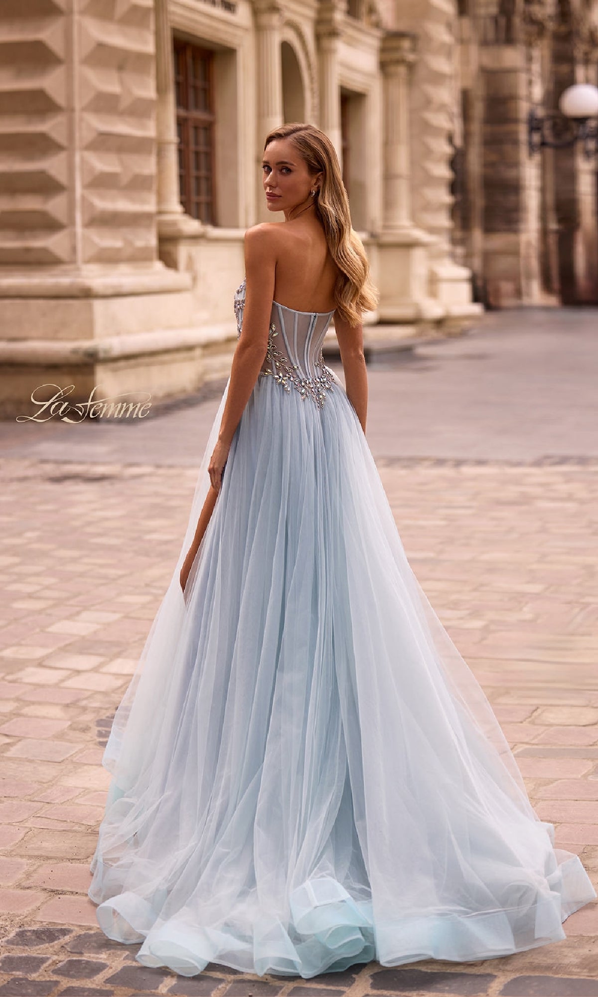 A woman in the La Femme 32862 Beaded-Corset Strapless Prom Gown with a sheer skirt stands on a cobblestone street by an ornate stone building, glancing over her shoulder.