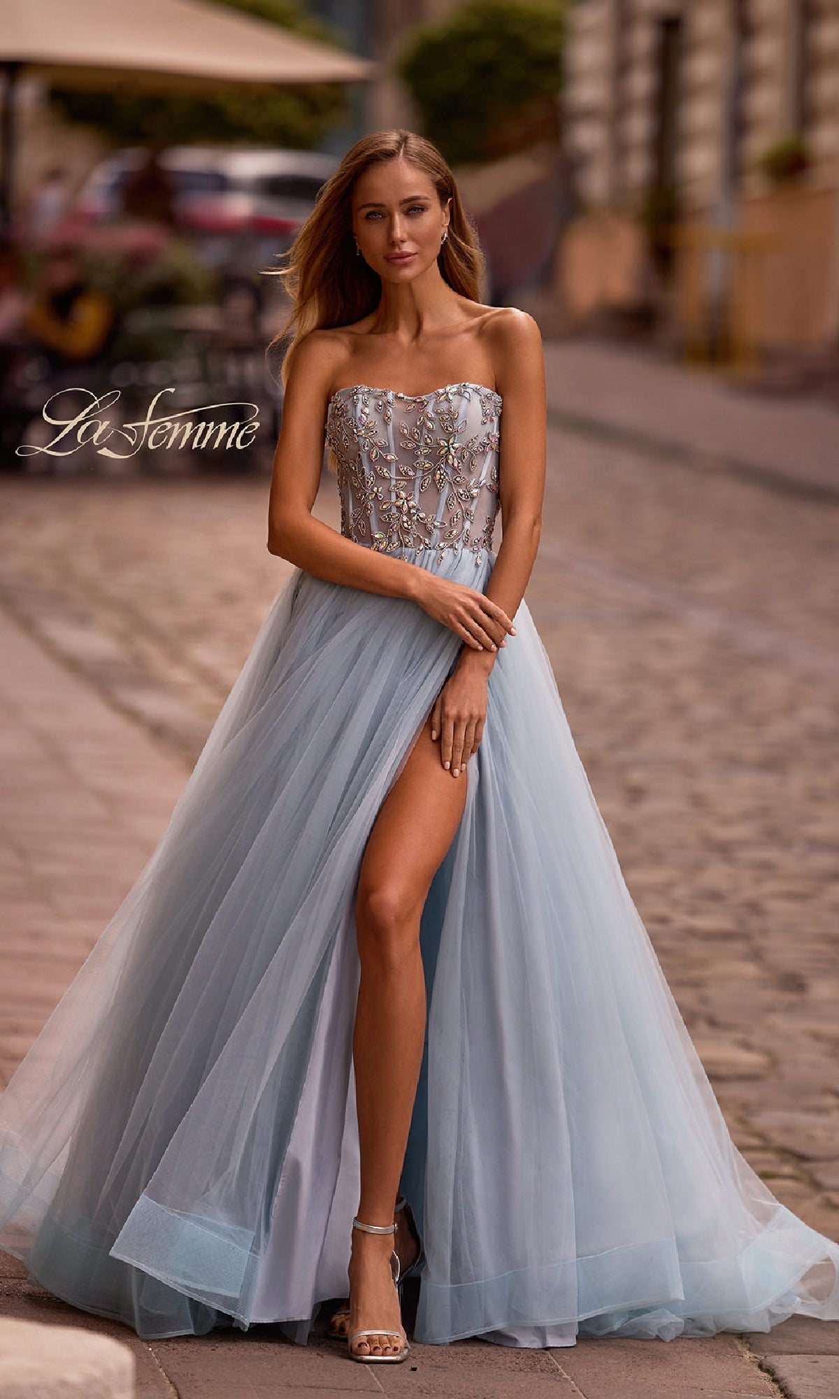 A woman in the La Femme 32862 Beaded-Corset Strapless Prom Gown with an a-line silhouette and thigh-high slit stands gracefully on a cobblestone street.