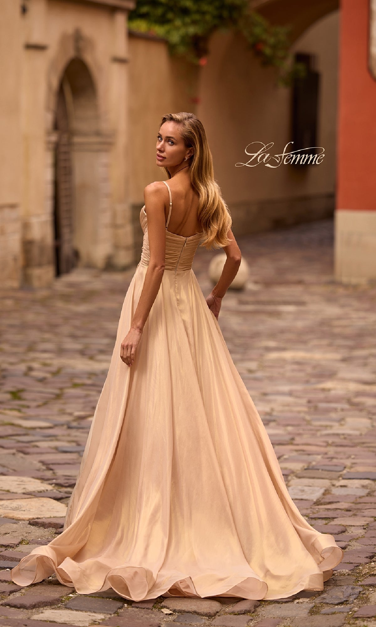 A woman in the Long Metallic Chiffon Prom Dress: La Femme 32864 stands on a cobblestone street, looking over her shoulder. The elegant a-line gown shimmers as it flows beautifully in the outdoor setting.