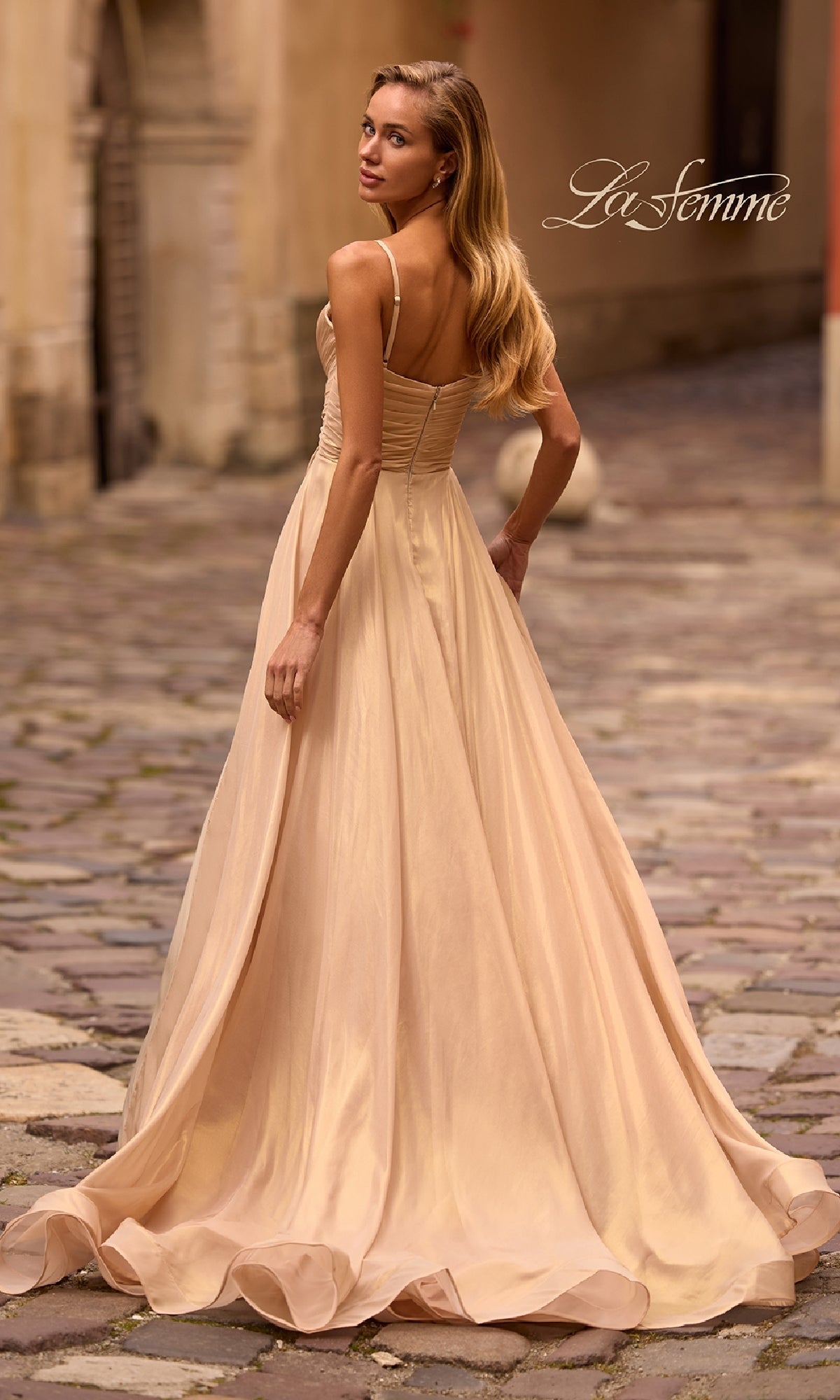 A woman in the La Femme 32864 Long Metallic Chiffon Prom Dress stands on a cobblestone street, glancing over her shoulder. The