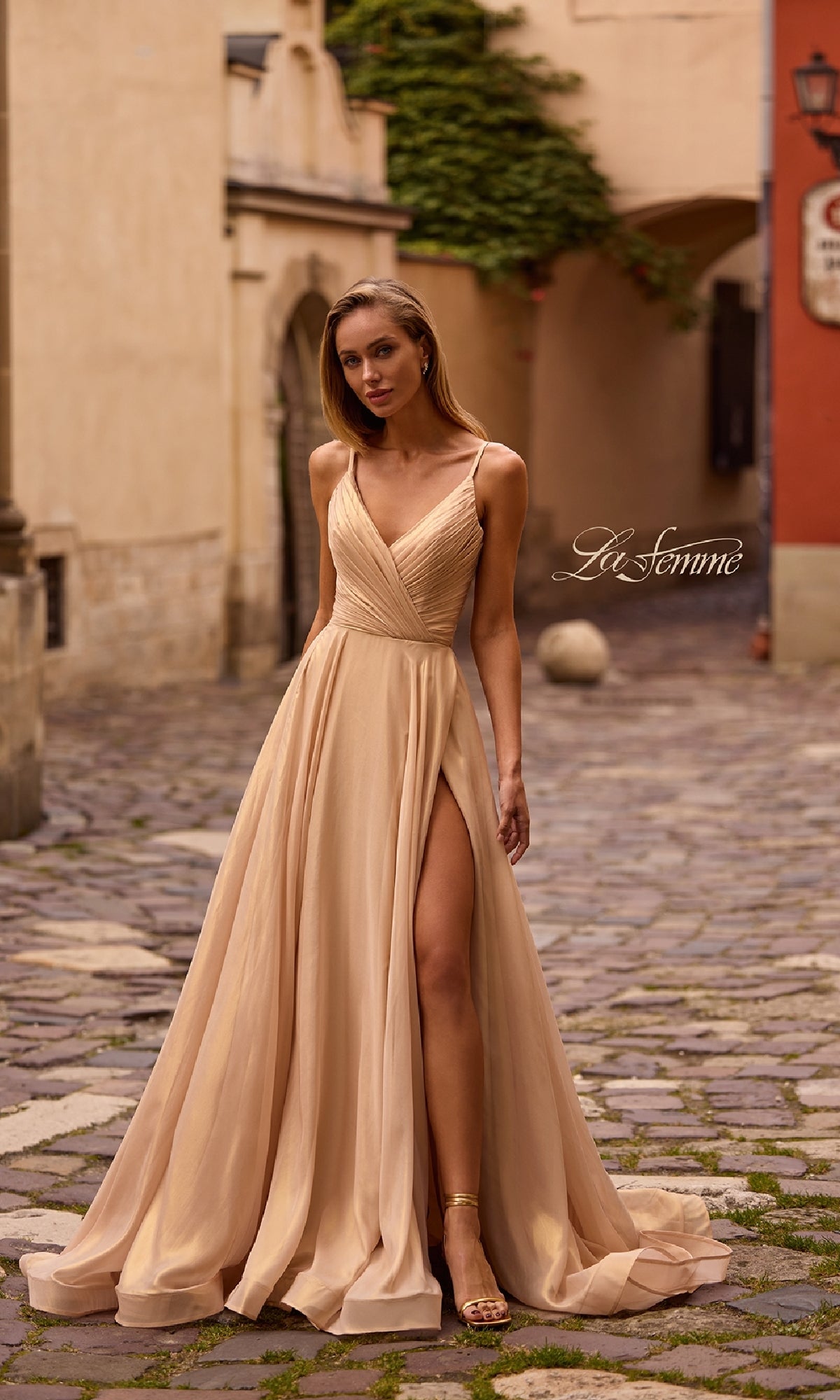 A woman poses on a cobblestone street wearing the La Femme 32864 Long Metallic Chiffon Prom Dress, a light beige gown with a flowing silhouette and thigh-high slit.