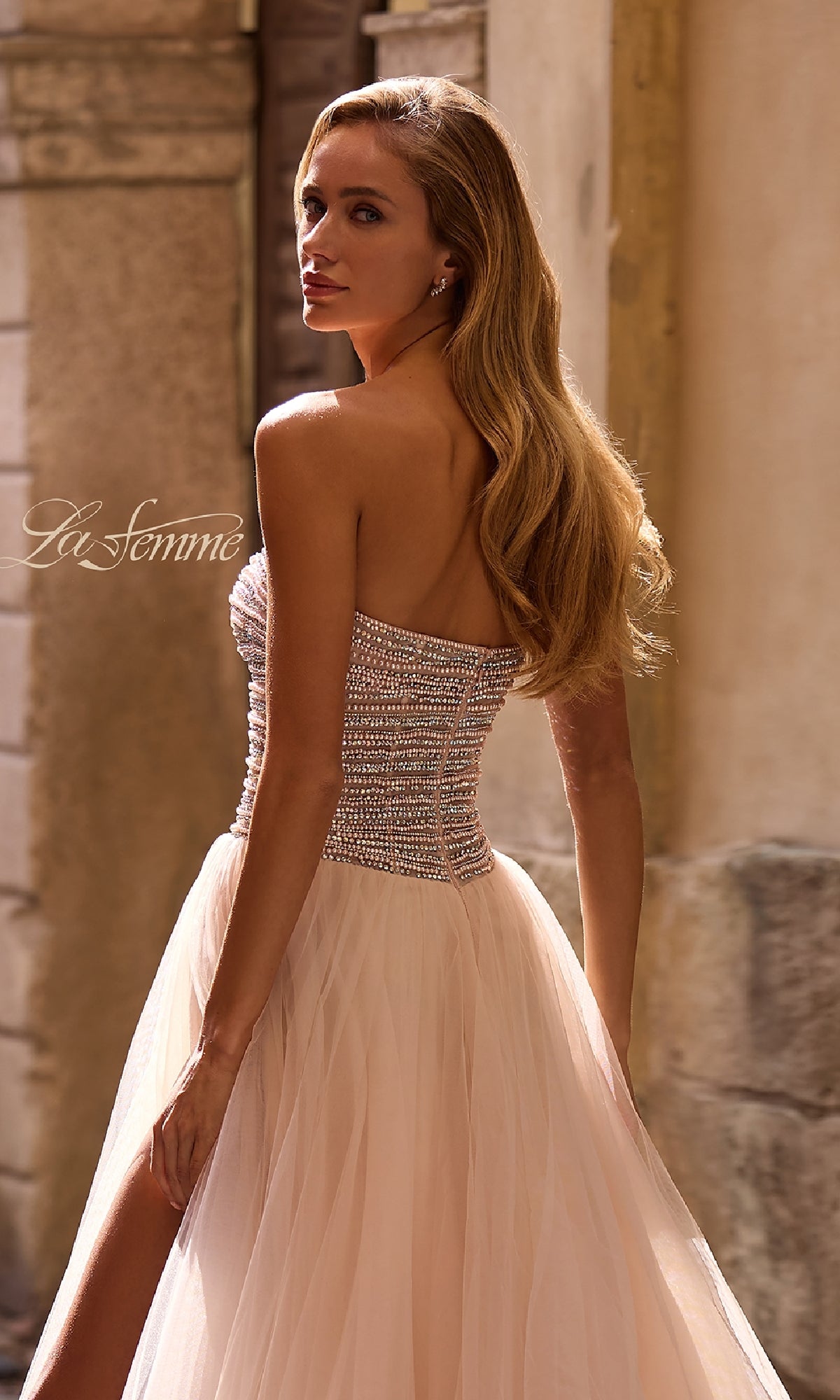 A woman in the Strapless Long Prom Ball Gown: La Femme 32875, featuring beaded embellishments and a tulle skirt, stands outdoors looking over her shoulder.