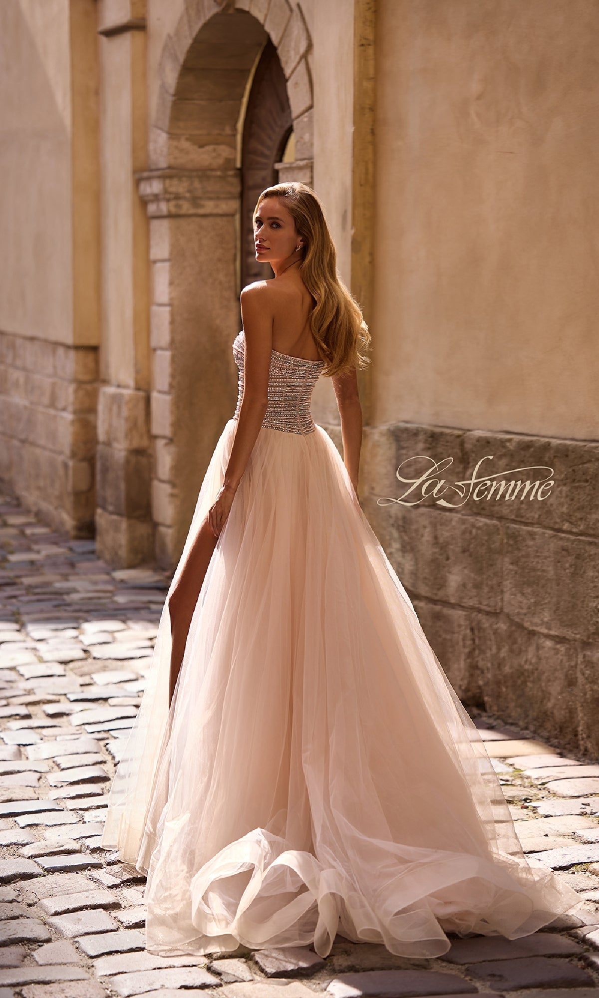 A woman wearing the Strapless Long Prom Ball Gown: La Femme 32875 with beaded details and a tulle skirt stands on a cobblestone street, glancing over her shoulder.