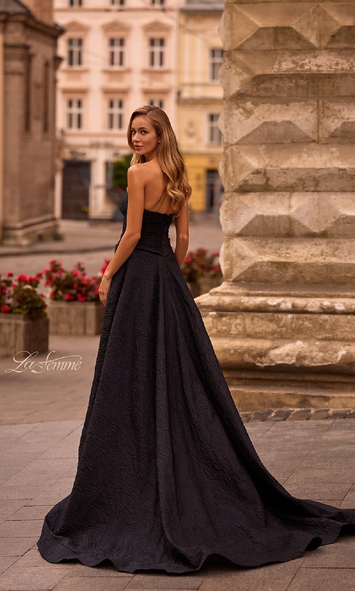 A woman wearing the Textured Strapless Black Prom Dress: La Femme 32893 stands outdoors on stone pavement, glancing over her shoulder with historic buildings in the background.