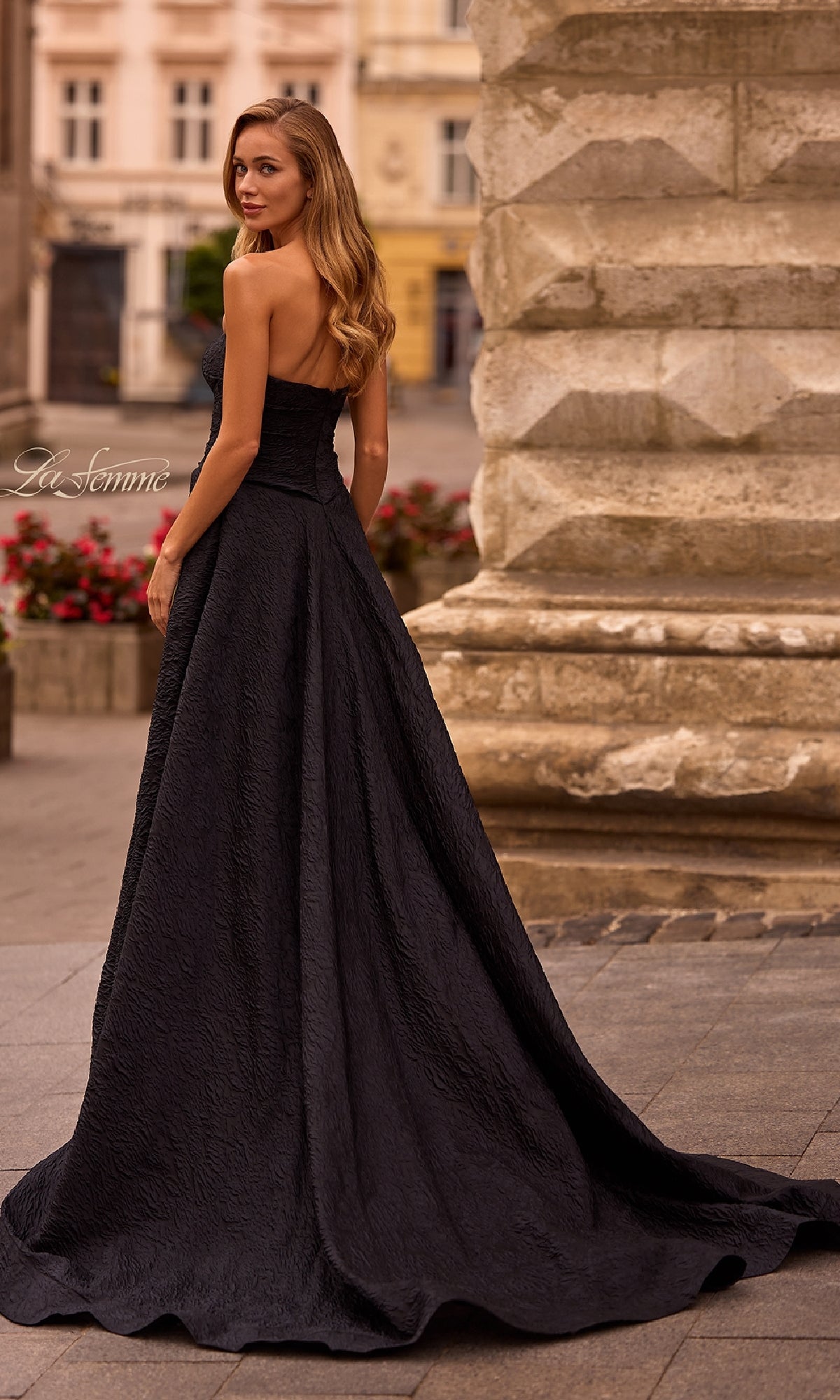 A woman in the La Femme 32893 Textured Strapless Black Prom Dress stands outdoors by a stone structure, glancing over her shoulder.