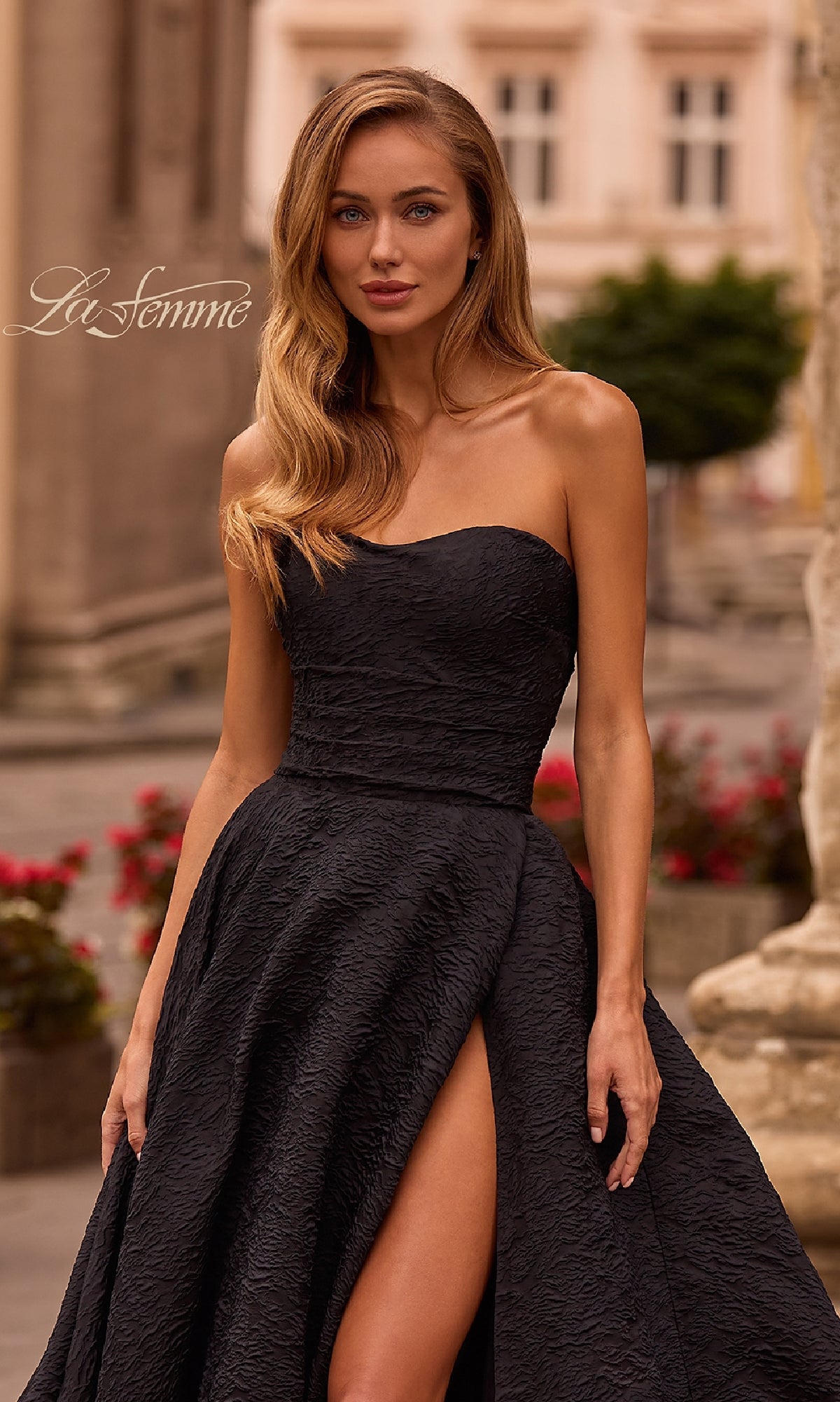 A woman stands outdoors amid historic architecture and blooming flowers, wearing the La Femme 32893 Textured Strapless Black Prom Dress with a high slit.