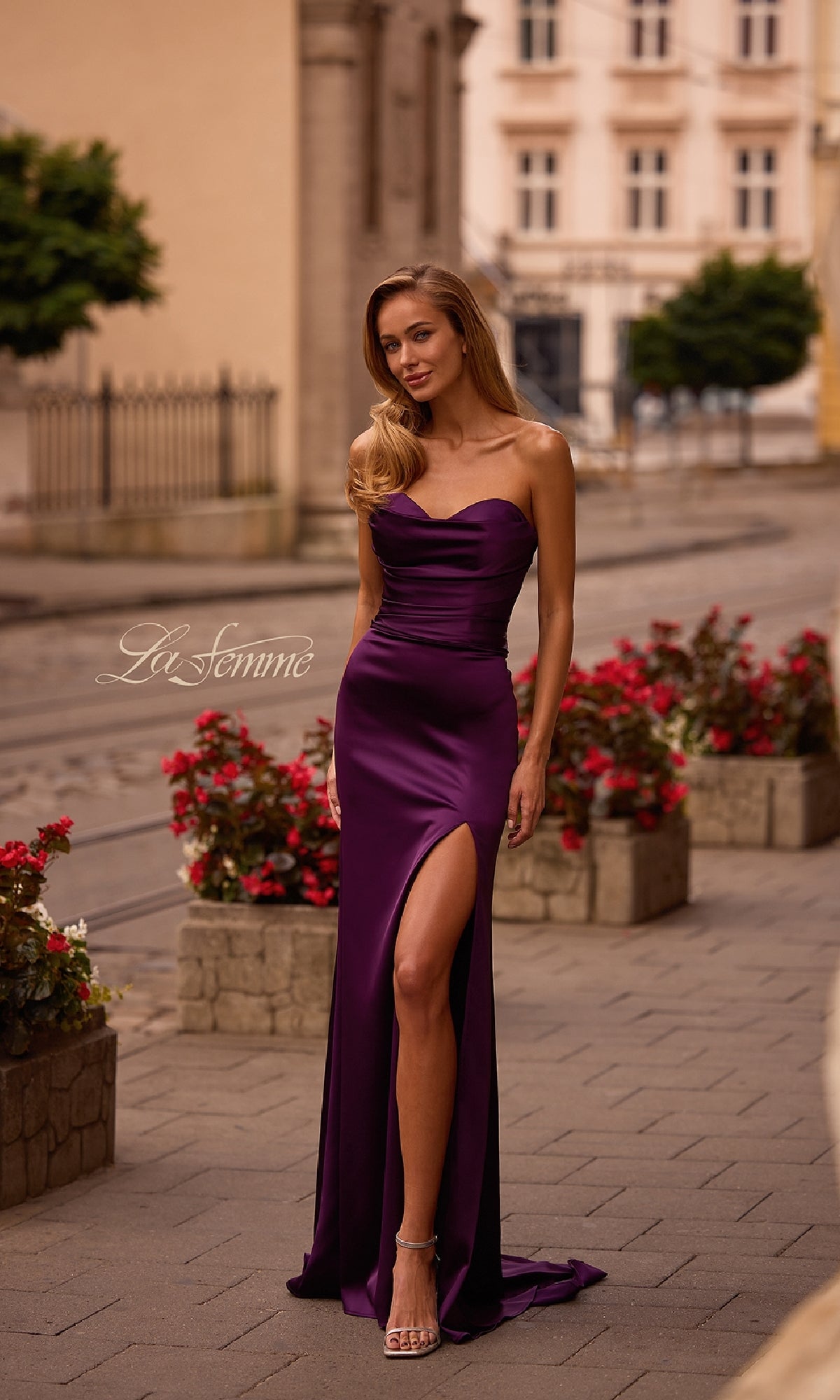 A woman stands outdoors on a paved street lined with planters and red flowers, wearing the La Femme 32896 Strapless Sweetheart Prom Gown in purple, featuring a long silhouette and thigh-high slit.