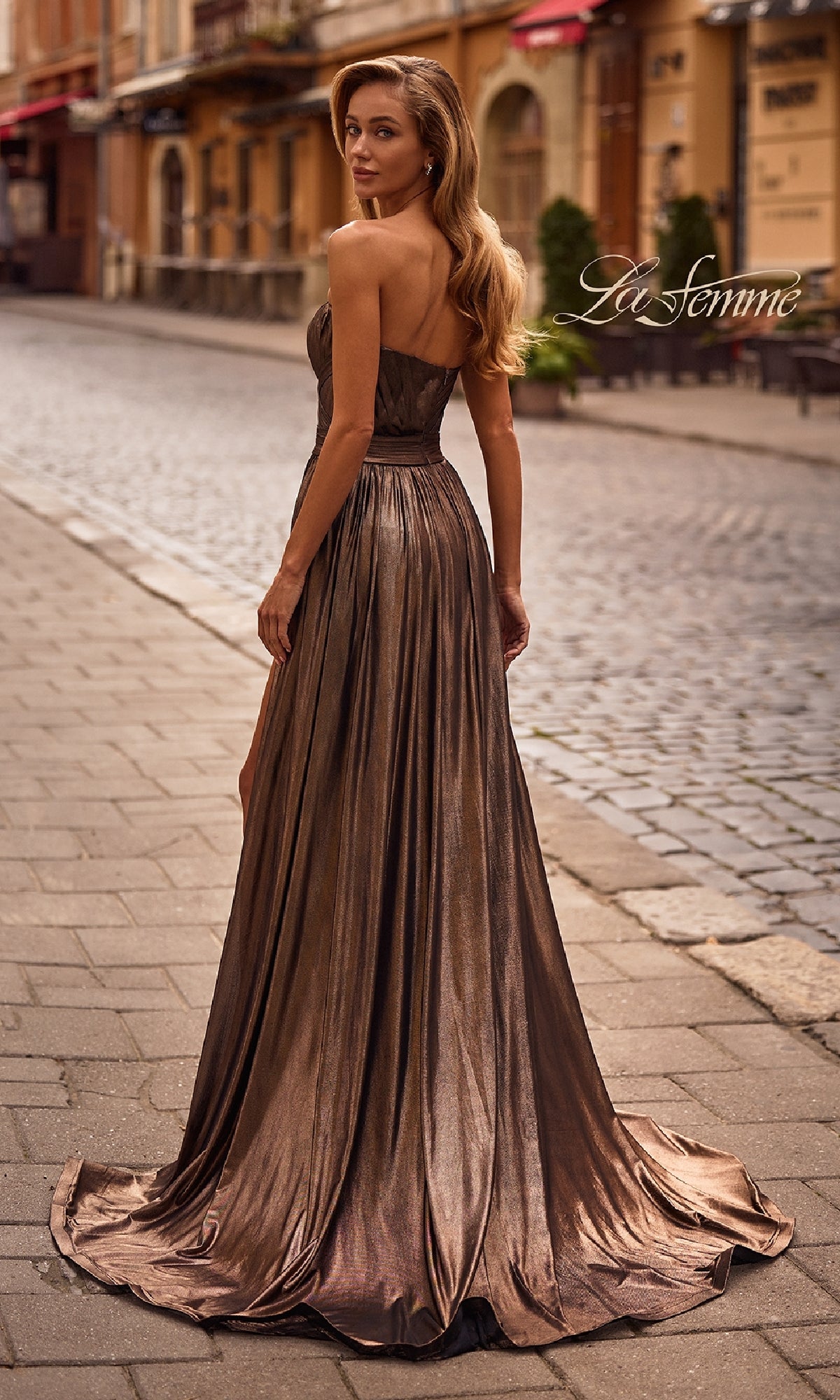 A woman stands on a cobblestone street, looking over her shoulder in the Strapless Long Metallic Prom Gown: La Femme 32988. The brand name